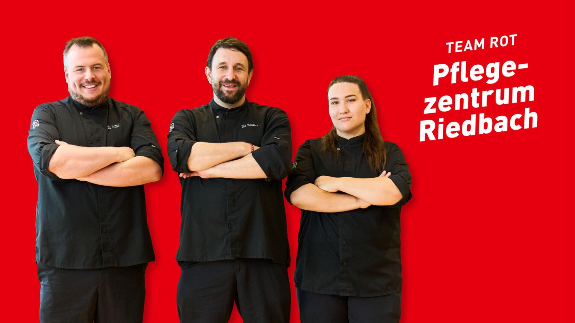 Drei Personen in schwarzen Kochuniformen stehen mit verschränkten Armen vor einem roten Hintergrund, auf dem der Schriftzug TEAM ROT Pflegezentrum Riedbach zu lesen ist.