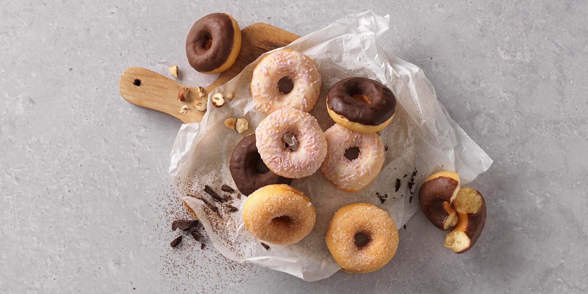 Verschiedene Donuts mit Schokolade, Zucker und rosa Zuckerguss sind auf einem Holzbrett und Backpapier angeordnet, umgeben von Krümeln und Haselnüssen auf einem grauen Untergrund.