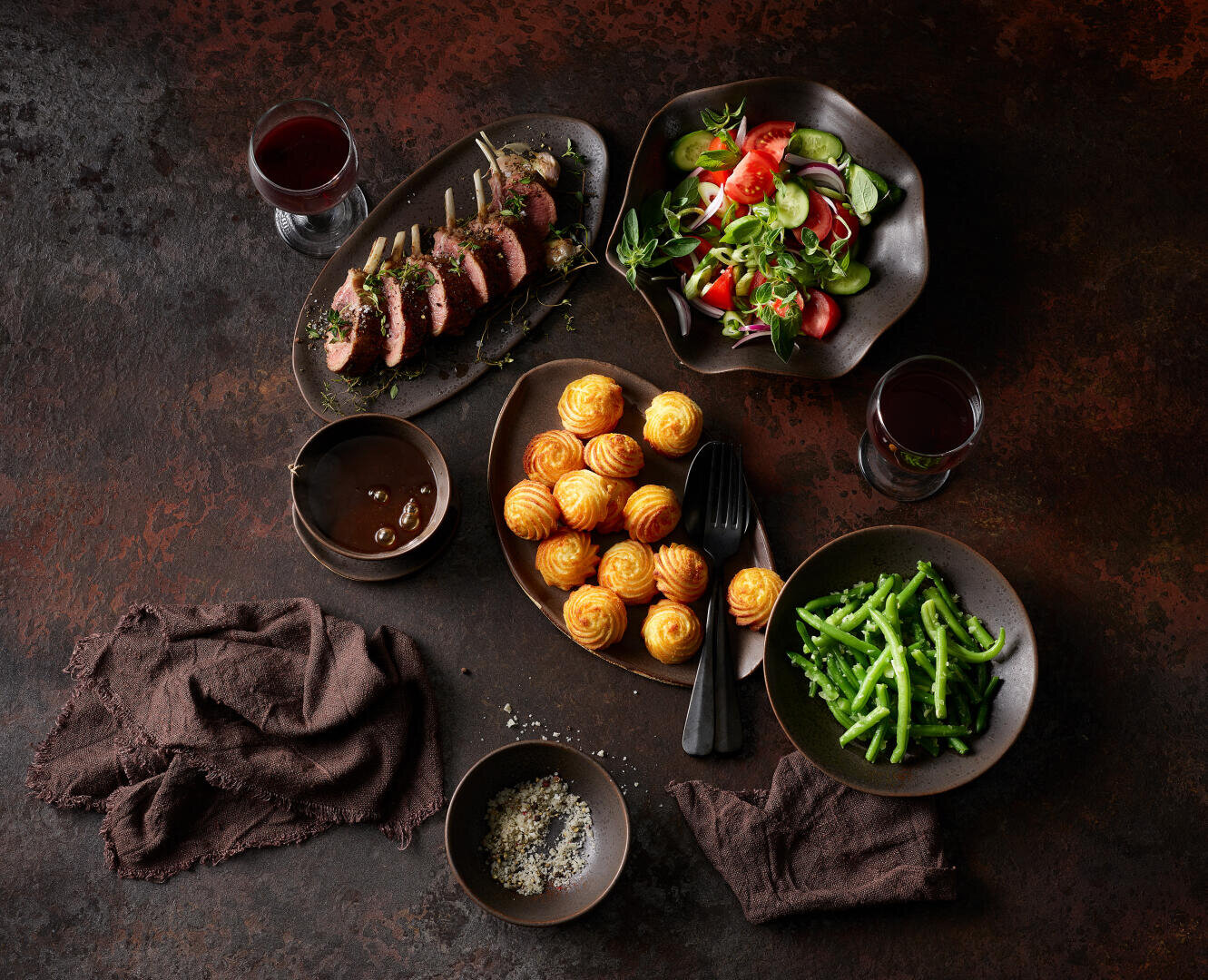 Ein elegantes Abendessen mit Lammscheiben, Salat, goldbraunen Kartoffelküchlein, grünen Bohnen, Rotwein, Soße sowie kleinen Schälchen mit Salz und Pfeffer, angerichtet auf einer dunklen, rustikalen Unterlage mit braunen Servietten und schwarzem Besteck.