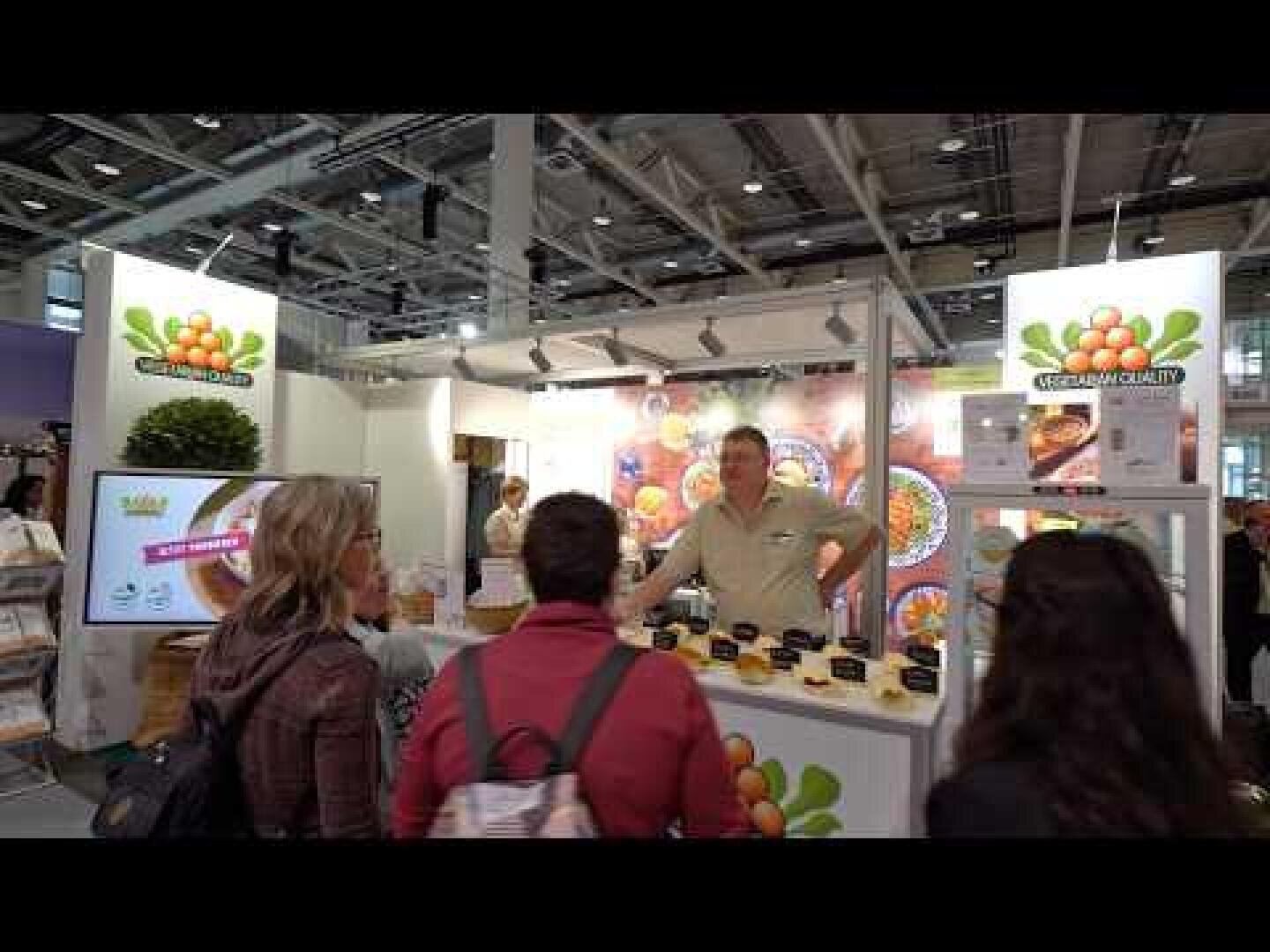 Drei Personen stehen an einem Messestand mit Schildern zum Thema Obst, während ein Verkäufer hinter der Theke in einer großen Messehalle verschiedene Produkte unter grellem Licht präsentiert.