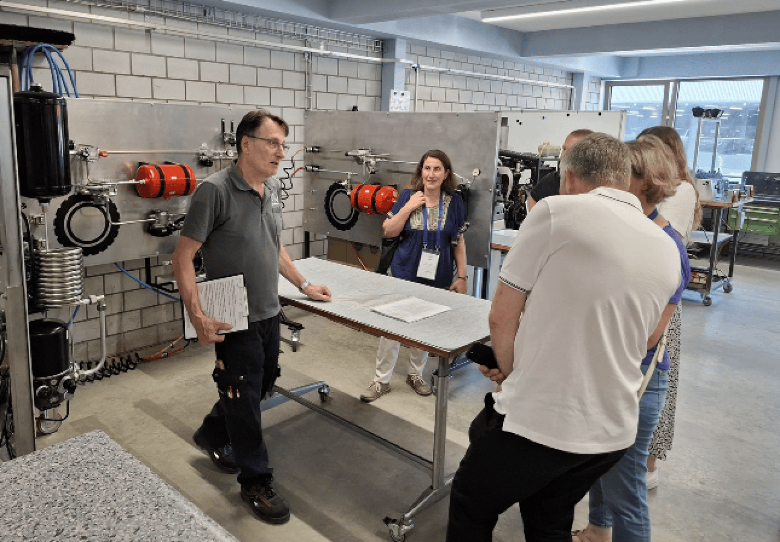 Ein Mann steht an einem Tisch und erklärt vier Erwachsenen in einer Werkstatt Maschinen. Hinter ihnen sind an einer Wand mechanische Geräte mit roten Tanks angebracht. Alle scheinen in ein Gespräch vertieft zu sein.