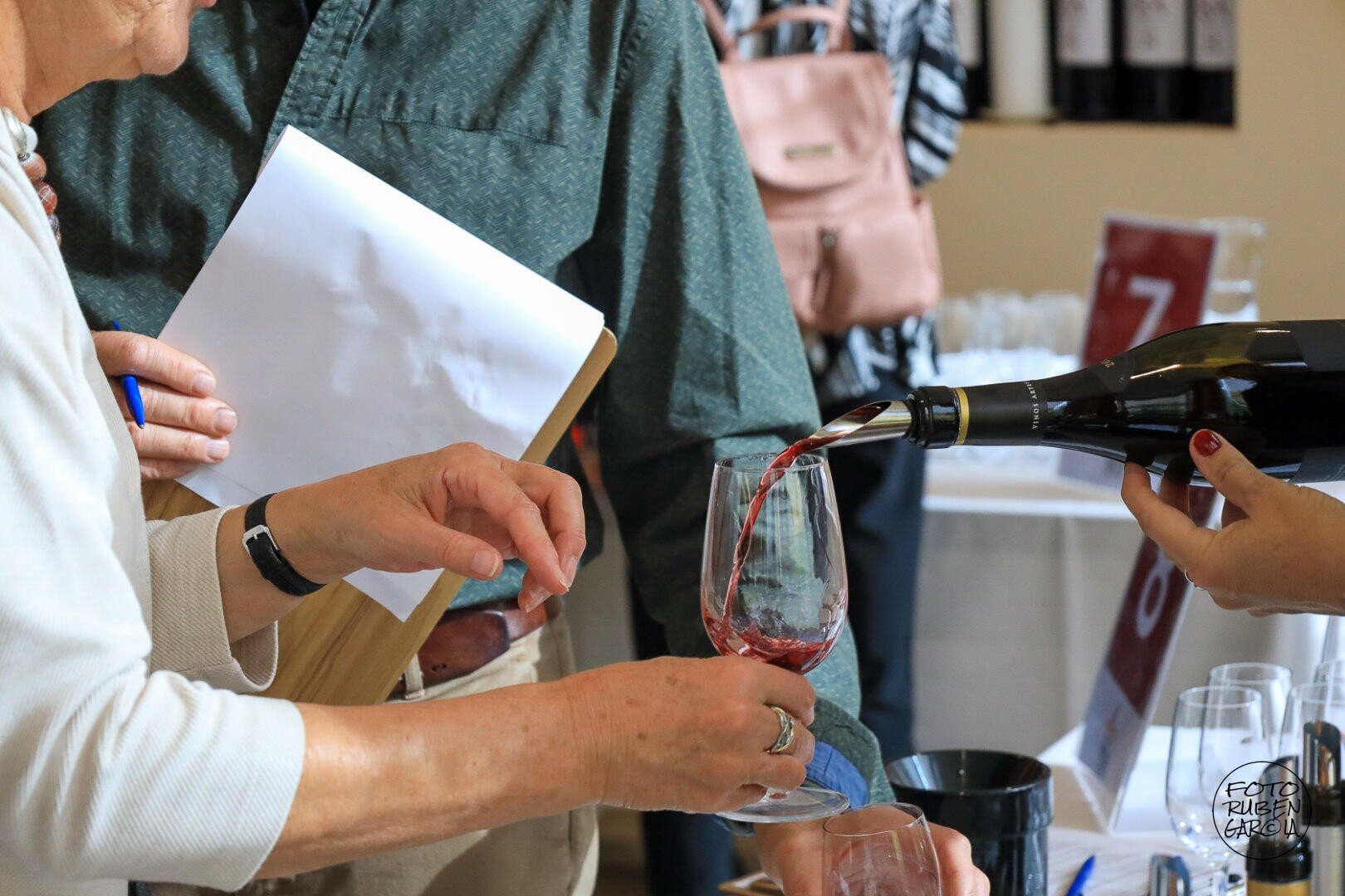 Eine Person schenkt Rotwein in ein Glas ein, das von einer anderen Person gehalten wird, während eine dritte Person in der Nähe ein Klemmbrett in der Hand hält. Im Hintergrund sind Flaschen, Gläser und nummerierte Schilder zu sehen – offenbar handelt es sich um eine Weinverkostung.