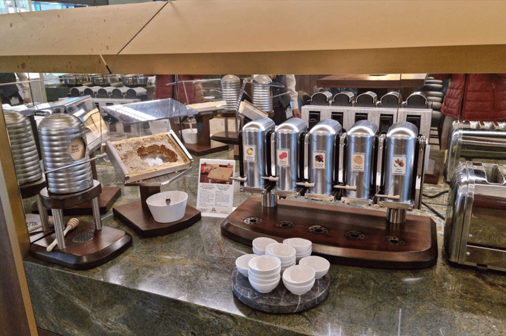 Buffetstation mit Waben, Honigspendern, kleinen Schälchen, Toastern und einem Stapel Teller auf einer grünen Marmorarbeitsplatte unter einer durchsichtigen Glas-Hustenschutzscheibe.