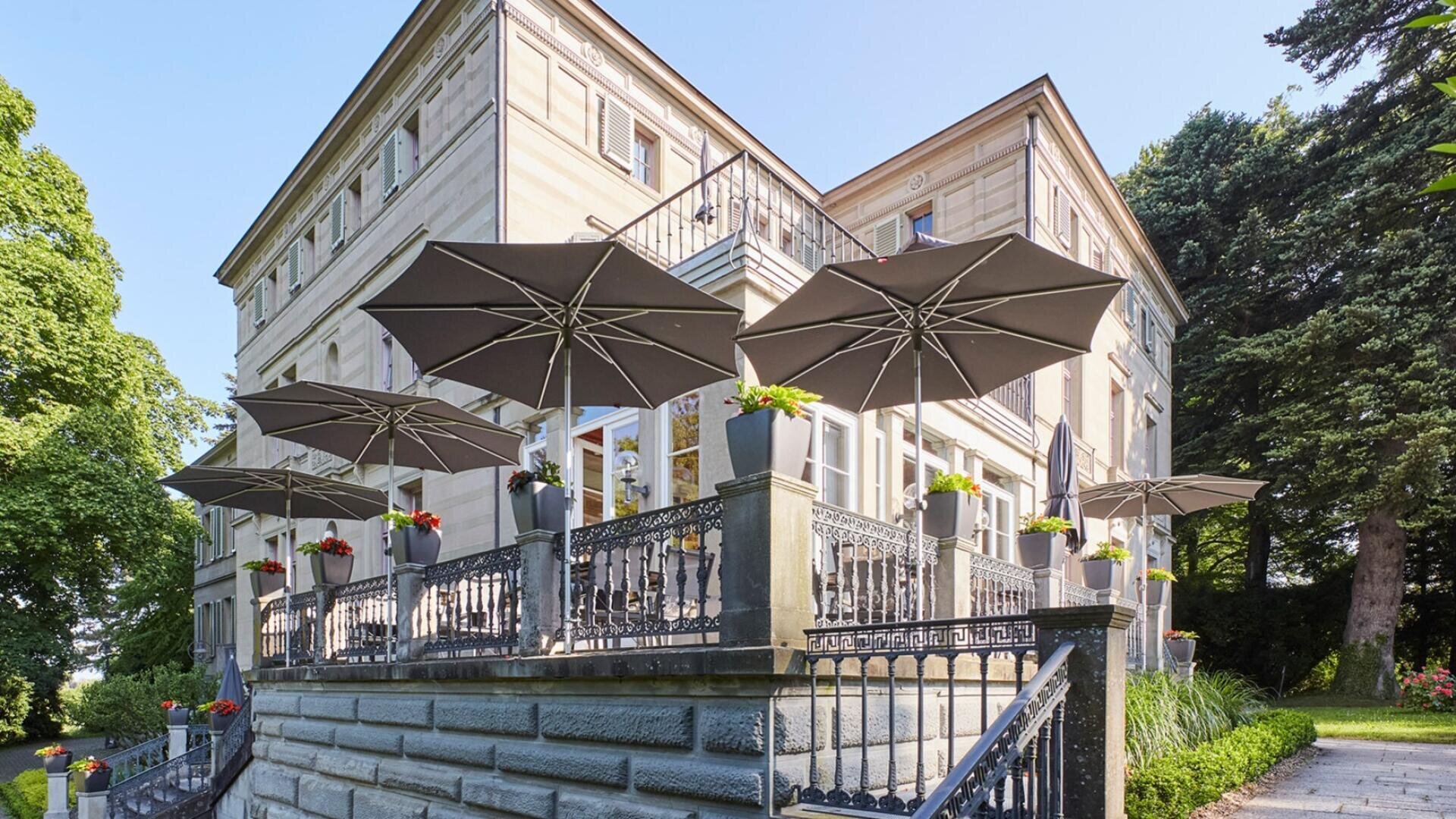 Historisches Steingebäude mit großen Fenstern und einer Terrasse. Schwarze Geländer und Blumen in Töpfen säumen die Terrasse, die von mehreren großen grauen Sonnenschirmen beschattet wird. An einem sonnigen Tag ist das Gebäude von Bäumen und Grün umgeben.
