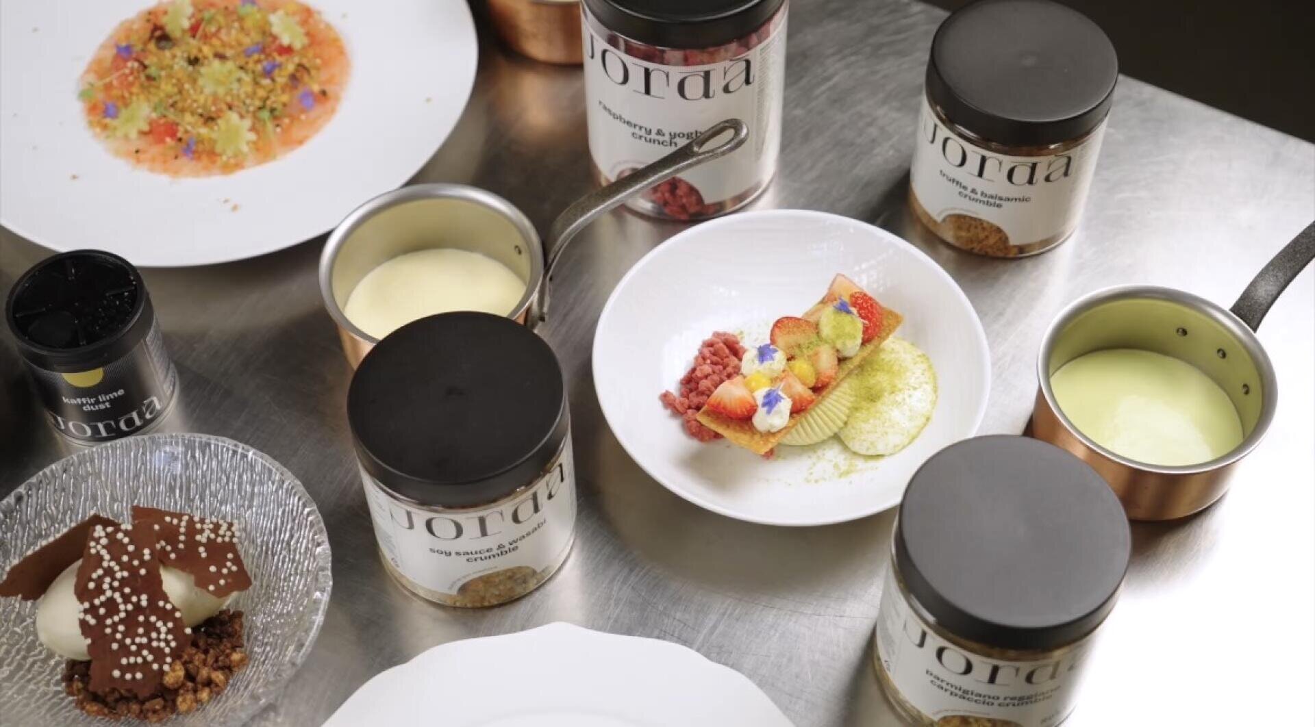 Auf einem Edelstahltisch sind Desserts und Gläser mit Zutaten angeordnet, darunter bunt angerichtete Süßspeisen, Pudding in Töpfen und Gläser mit der Aufschrift „Jorda“. Einige Gerichte sind mit Obst und essbaren Blüten garniert.
