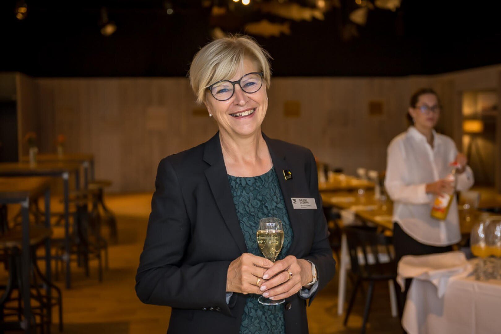 Eine lächelnde Frau in einem Blazer, die ein Weinglas in der Hand hält, steht in einem modernen Essbereich, während im Hintergrund eine unscharf abgebildete Person den Tisch deckt.
