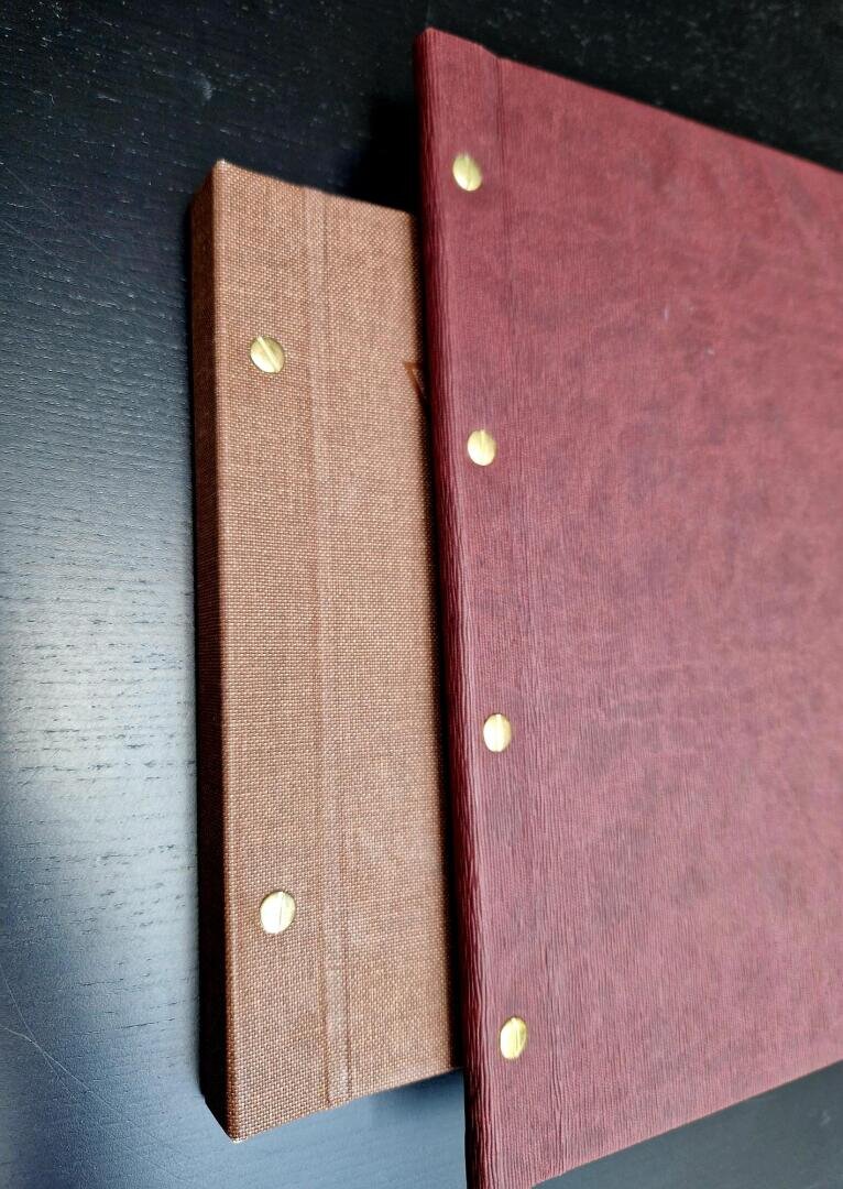 Zwei gebundene Bücher mit strukturierten Einbänden und goldenen Einbandschrauben liegen auf einer schwarzen Unterlage gestapelt, wobei das braune Buch teilweise von dem größeren roten Buch verdeckt wird.