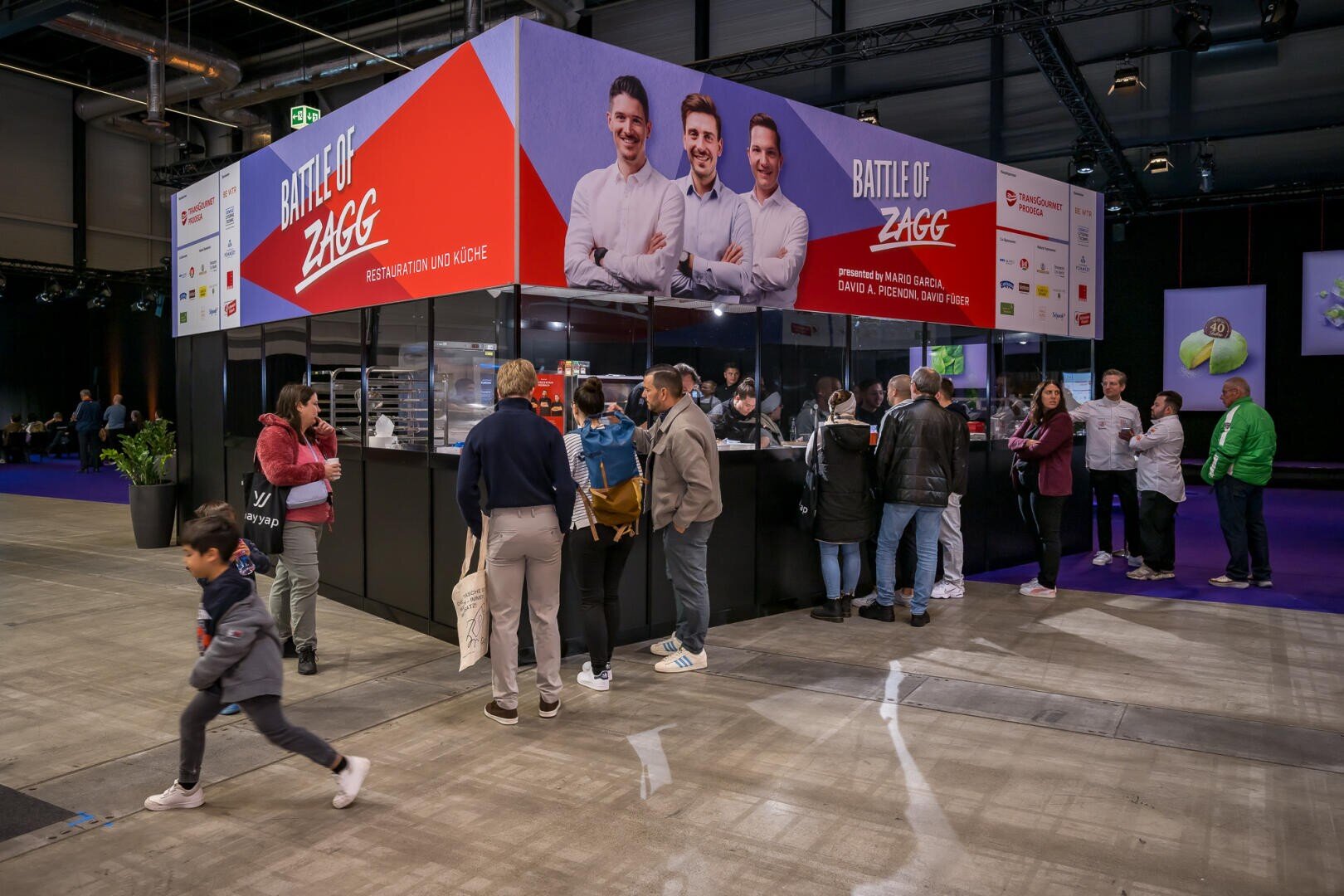 An einem Messestand mit dem Titel Battle of ZAGG ist ein großes Banner zu sehen, auf dem Fotos von drei Köchen und einer Gruppe von Menschen zu sehen sind, die in einer Schlange auf Essen warten.