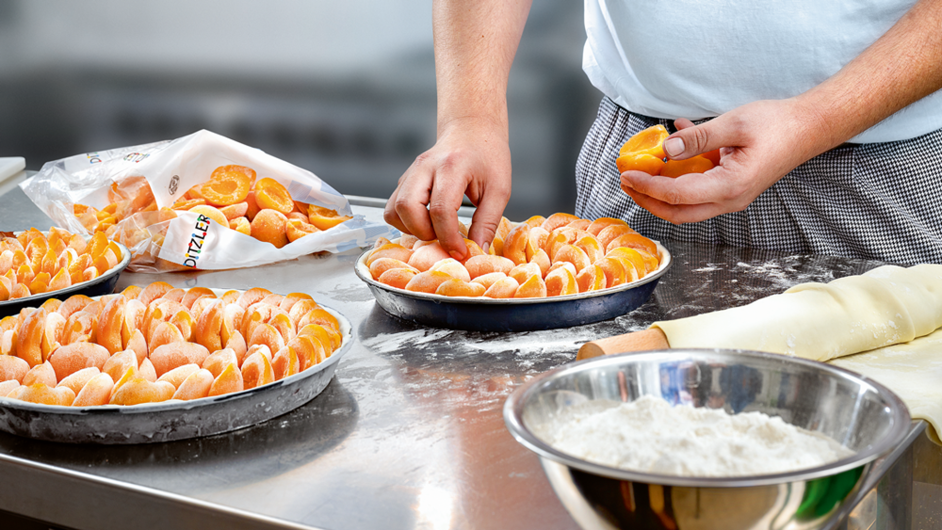 Eine Person ordnet Orangensegmente in einer Kuchenform auf einer Metallarbeitsplatte an, umgeben von weiteren Kuchen, einer Schüssel mit Mehl, einem Nudelholz und einer Tüte mit geschälten Orangenscheiben.