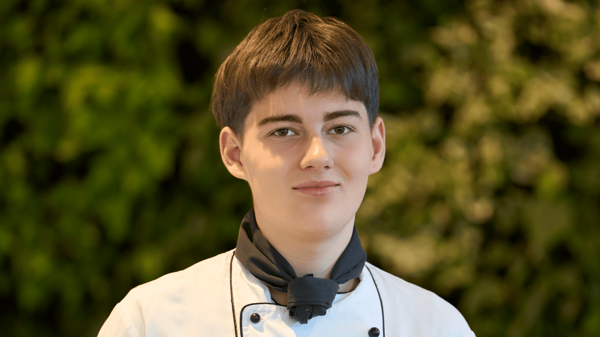 Ein junger Koch in weißer Uniform und mit schwarzem Halstuch blickt in die Kamera.