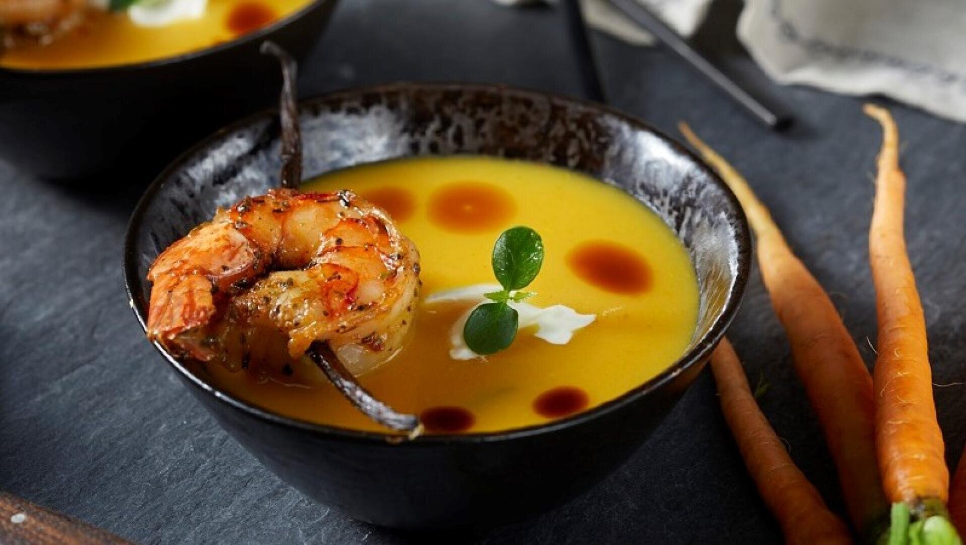 Eine Schüssel cremige Orangensuppe, garniert mit einem Sahnehauch, zwei gegrillten Garnelen am Spieß, kleinen Kräuterblättern und einigen Tropfen Soße, dazu frische Möhren neben der Schüssel.