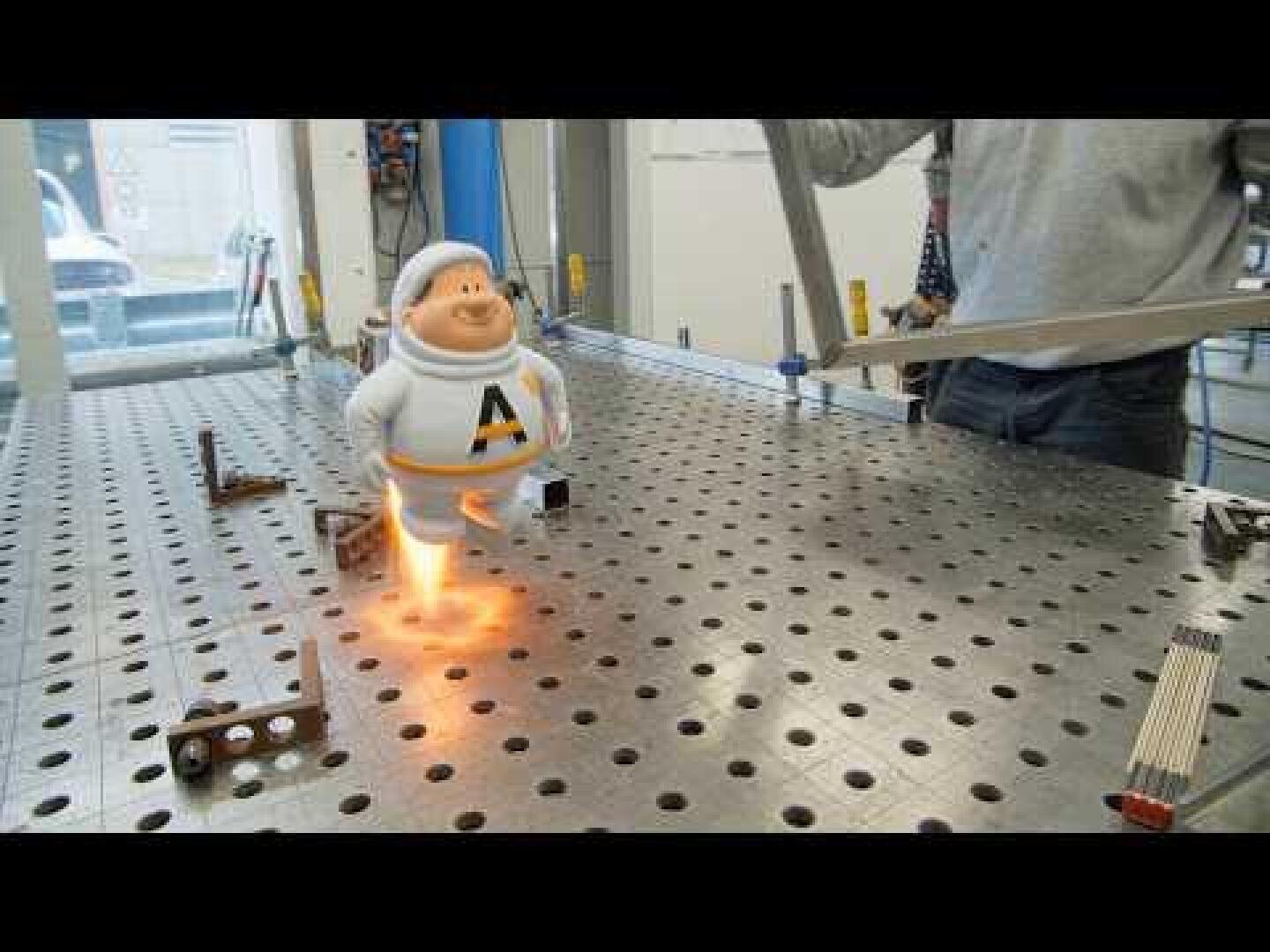 Ein kleines Astronauten-Spielzeug scheint gerade von einem perforierten Metalltisch abzustarten, mit Flammen unter seinen Füßen, während eine Person in der Nähe etwas einstellt.
