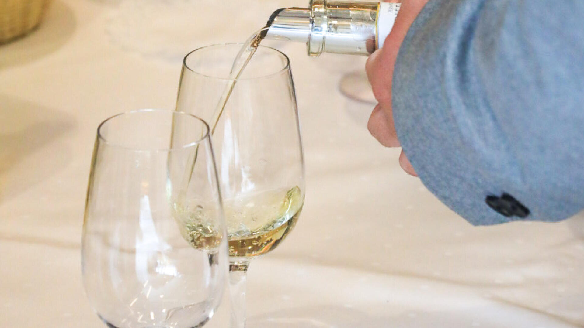 Eine Person mit einem blauen Ärmel schenkt Weißwein aus einer Flasche in eines von zwei leeren Weingläsern auf einer weißen Tischdecke ein.