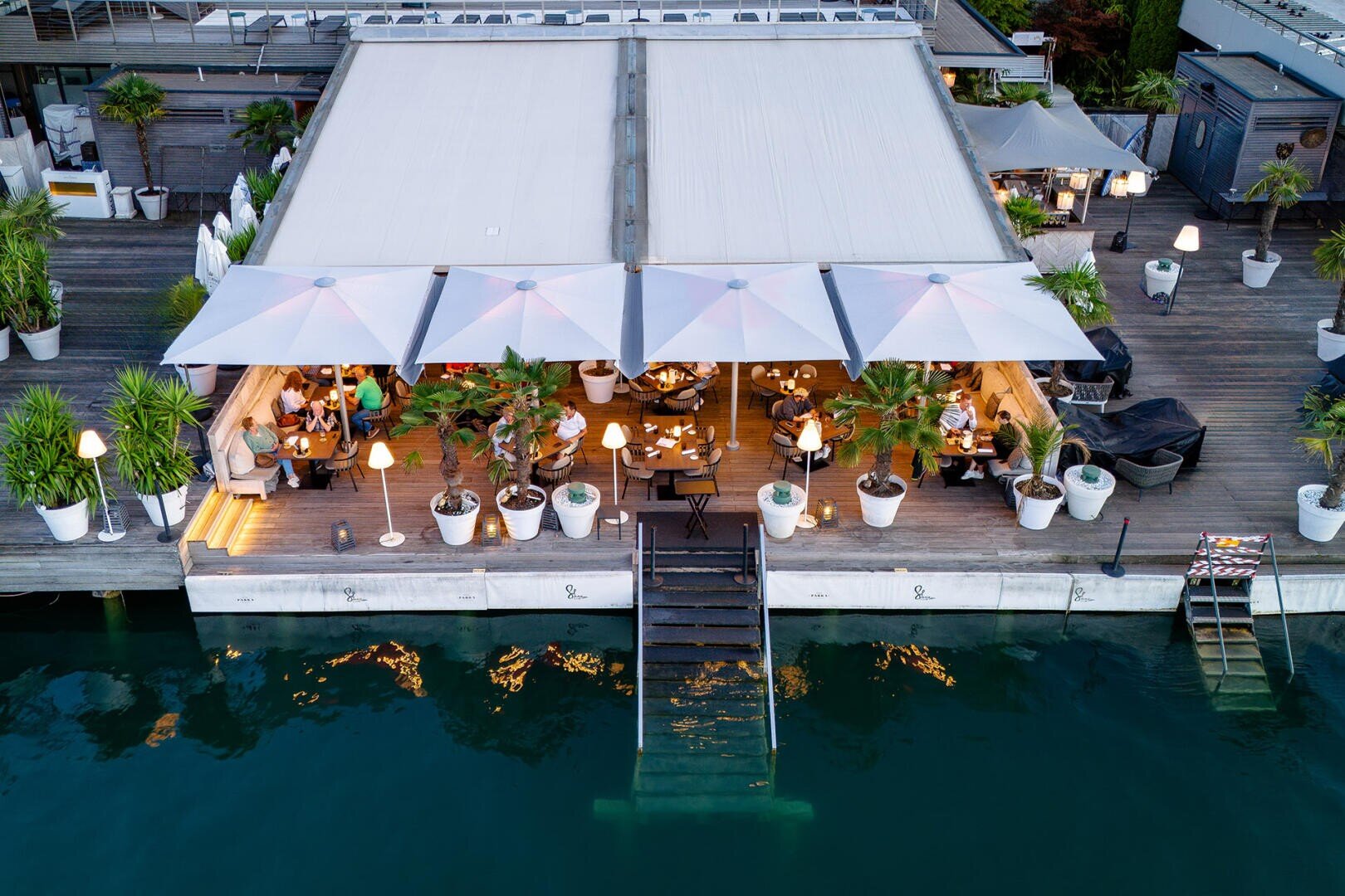 Luftaufnahme eines Restaurants am Wasser mit weißen Sonnenschirmen, Sitzgelegenheiten im Freien, Topfpflanzen und Lichtern, die sich im Wasser spiegeln. Eine Treppe führt von der Terrasse hinunter zum Wasser.