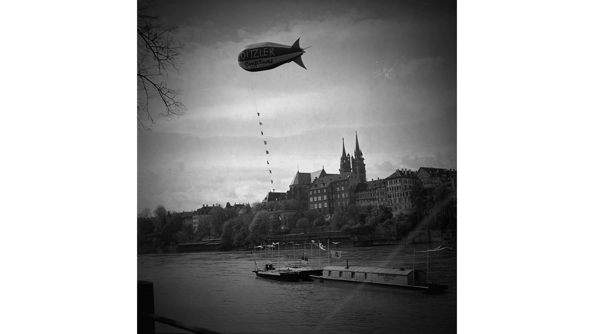 Ein Schwarz-Weiß-Foto eines Flusses mit Booten und einem schwimmenden Luftschiff, das für OZZLER wirbt. Im Hintergrund sind historische Gebäude mit Türmen unter einem bewölkten Himmel zu sehen.