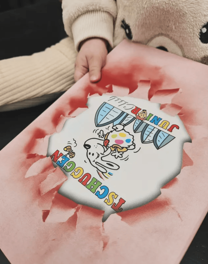 Eine Kinderhand hält eine rosa Mappe mit einem eingerissenen Verschluss, durch den eine bunte TSCHUGGEN JUNIOR-Cartoon-Illustration mit einem Hund und darunter liegenden Gebäuden zu sehen ist. Der Hintergrund ist schwarz.