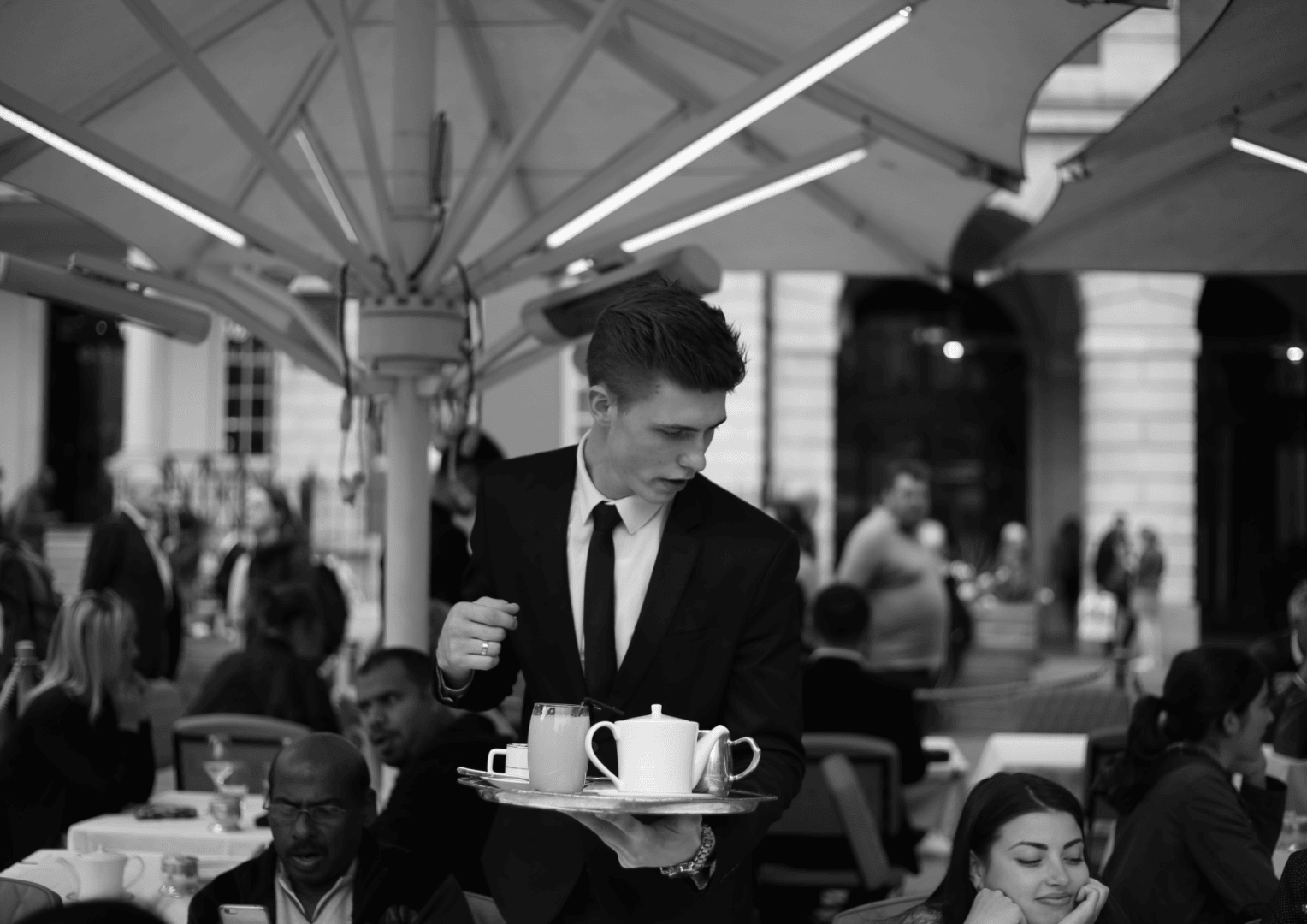 Ein Kellner im Anzug trägt in einem Straßencafé ein Tablett mit einer Teekanne und Tassen. An den Tischen sitzen Menschen und unterhalten sich, während große Sonnenschirme für Schatten sorgen. Im Hintergrund sind Säulen und Torbögen zu sehen.