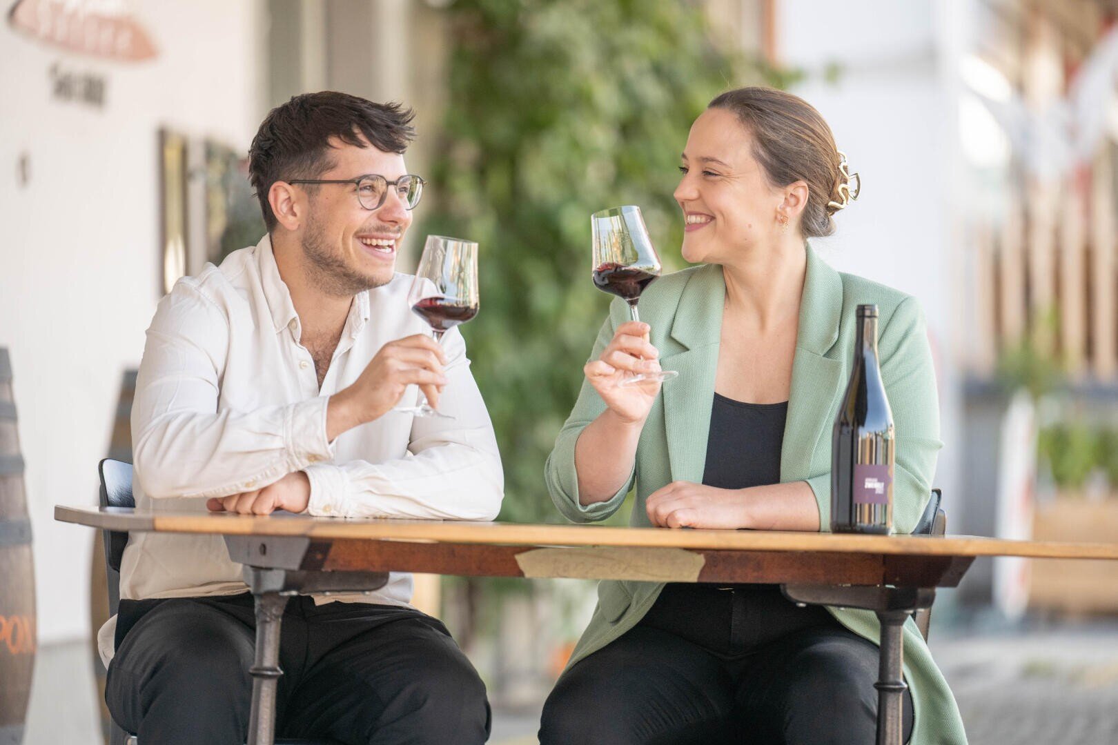 Zwei Menschen sitzen an einem Tisch im Freien, lächeln und stoßen mit ihren Gläsern Rotwein an. Auf dem Tisch steht eine Weinflasche, und die Atmosphäre wirkt entspannt und ungezwungen, mit viel Grün im Hintergrund.