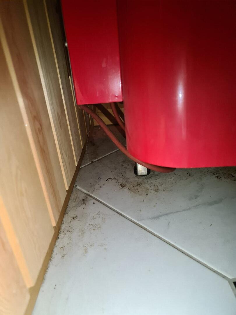 Eine Nahaufnahme einer staubigen Bodenecke mit weißen Fliesen, einem roten Gerät oder Gegenstand und einer Wand aus Holzpaneelen.