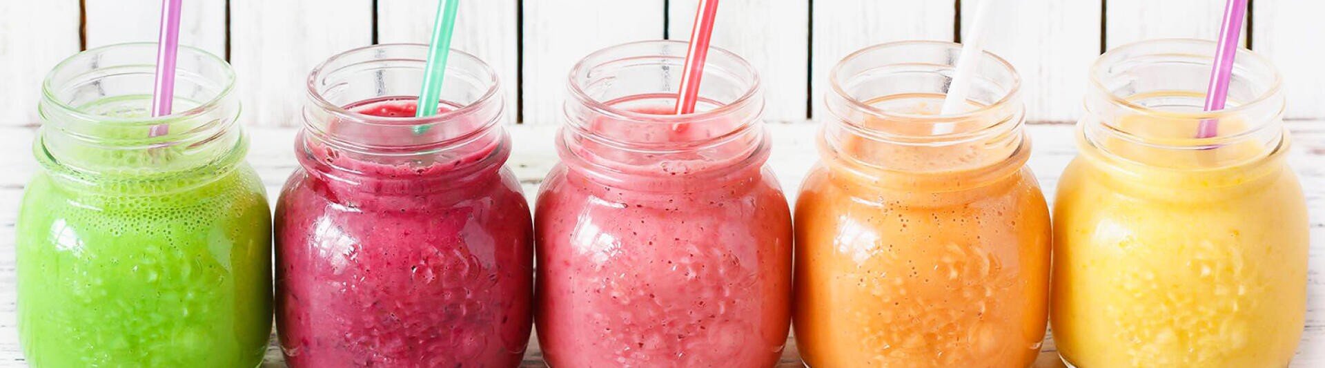 Fünf Einmachgläser, gefüllt mit bunten Smoothies – grün, lila, rosa, orange und gelb –, jedes mit einem passenden Strohhalm, standen auf einer weißen Holzplatte in einer Reihe.