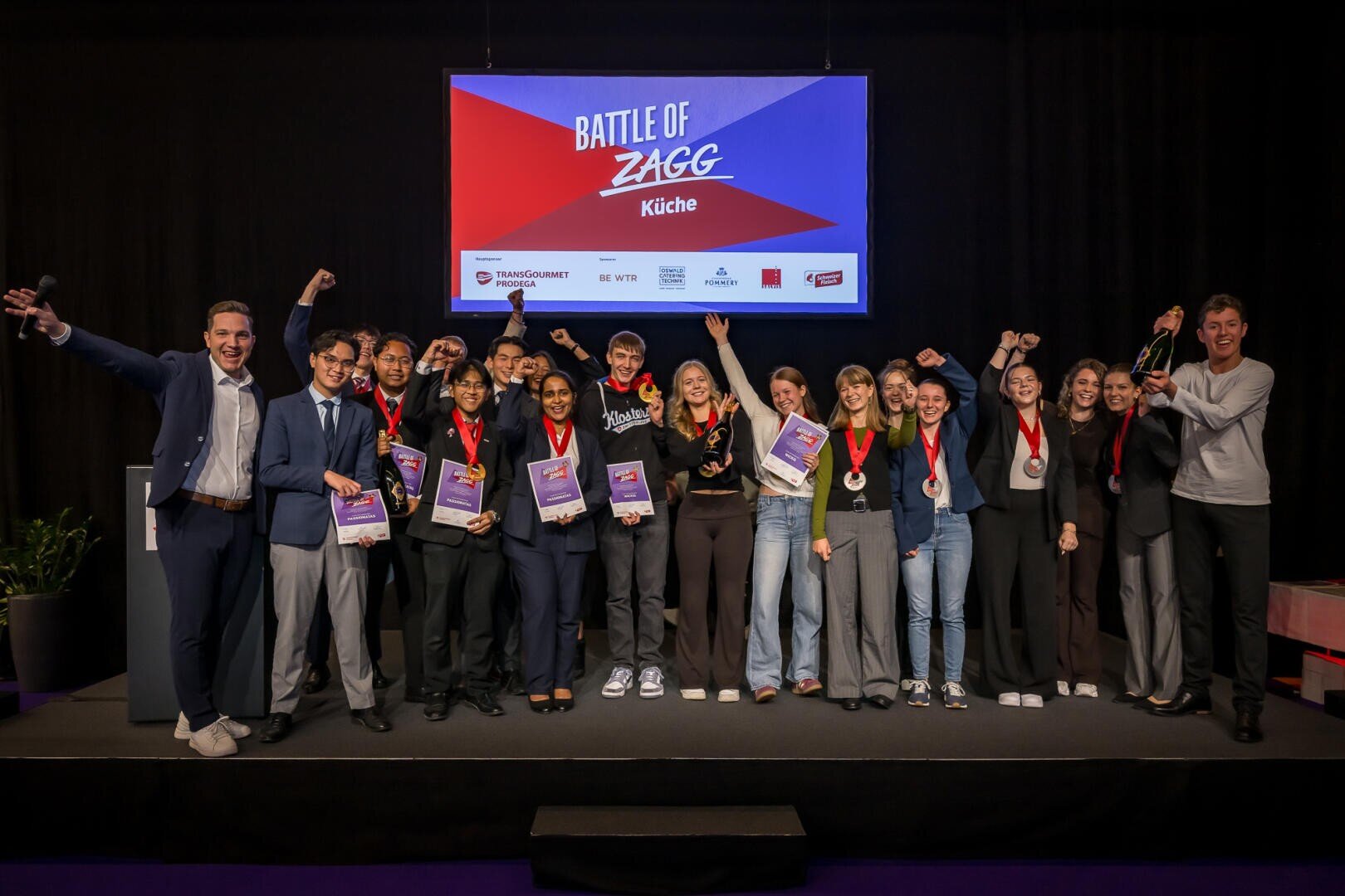 Eine Gruppe von Preisträgern steht auf einer Bühne vor einer Leinwand mit der Aufschrift Battle of Zagg Küche; viele von ihnen halten Urkunden und Medaillen in den Händen.