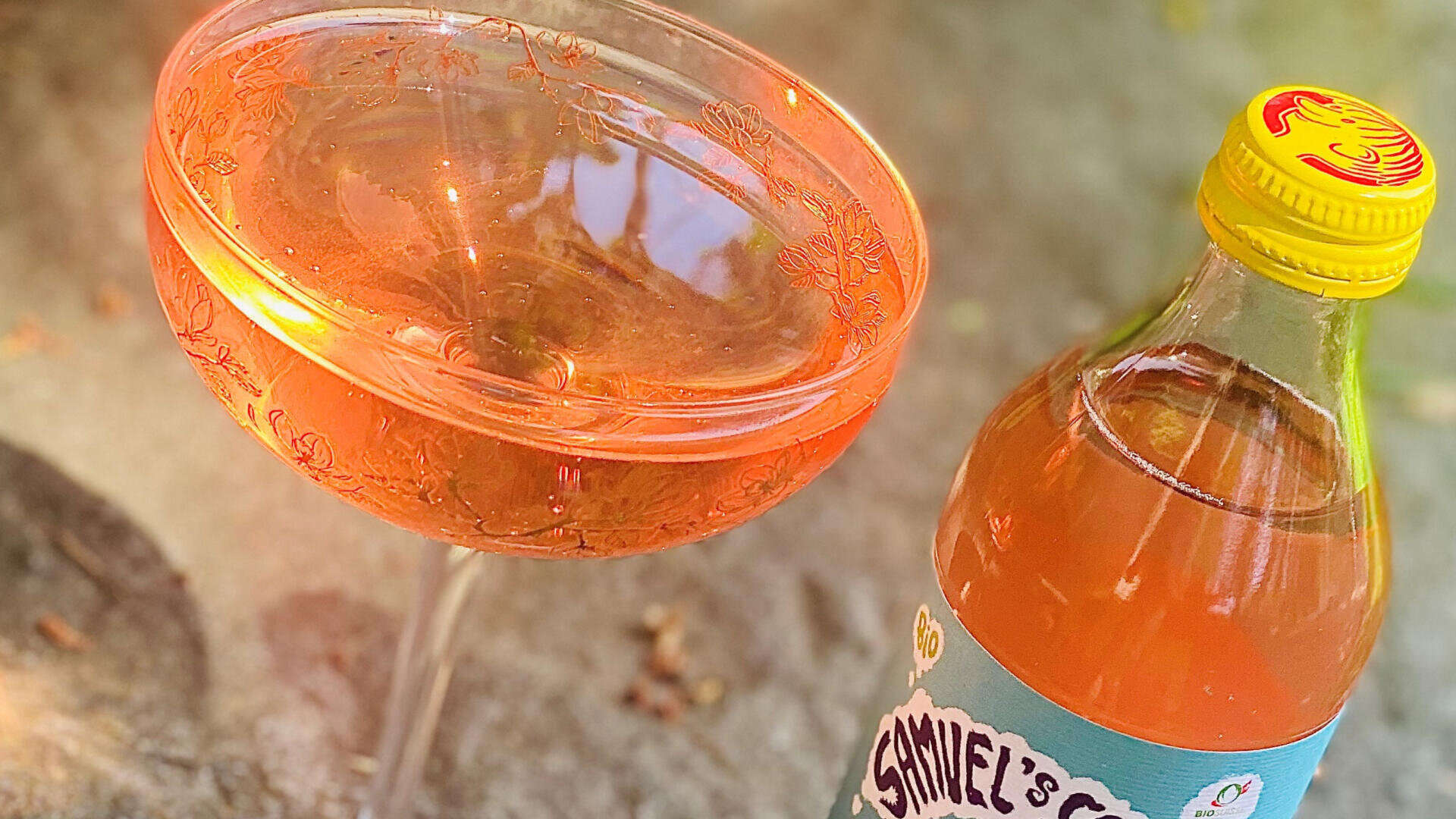 Ein Coupé-Glas, gefüllt mit einem rosa kohlensäurehaltigen Getränk, steht neben einer Flasche mit der Aufschrift „Samuels Cider“, die eine orangefarbene Flüssigkeit enthält und einen gelben Verschluss hat, auf einer Steinoberfläche im Sonnenlicht.