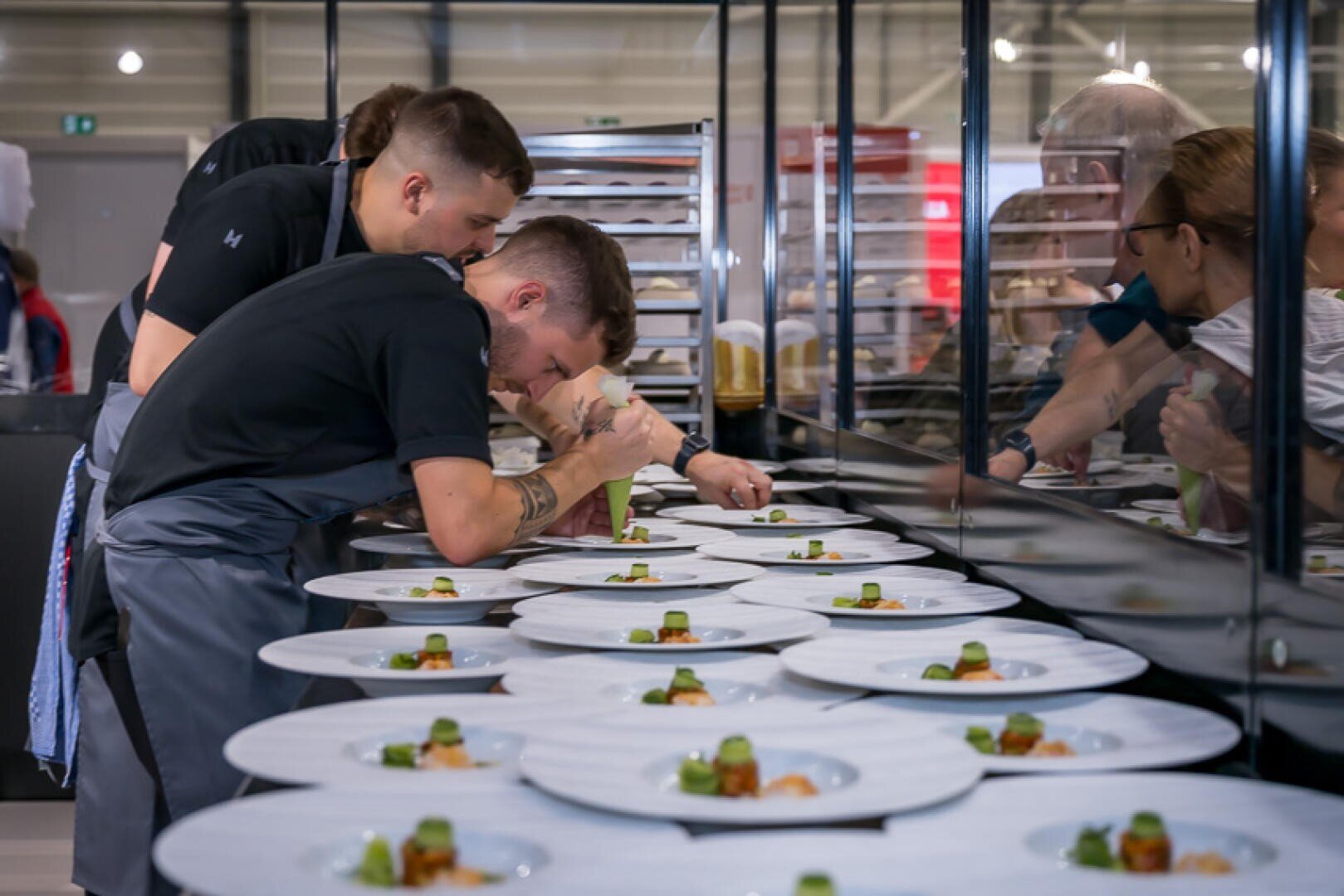 Köche in schwarzen Hemden und grauen Schürzen konzentrieren sich in einer professionellen Küche darauf, Gourmetgerichte anzurichten, und ordnen die Speisen sorgfältig auf mehreren weißen Tellern an, die auf einer Arbeitsplatte aufgereiht sind.