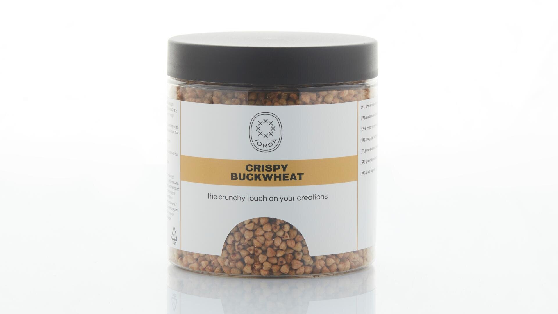 Ein durchsichtiges Plastikglas mit schwarzem Deckel, gefüllt mit knusprigem Buchweizen. Auf dem weiß-gelben Etikett steht „CRISPY BUCKWHEAT“ und „die knusprige Note für Ihre Kreationen“.
