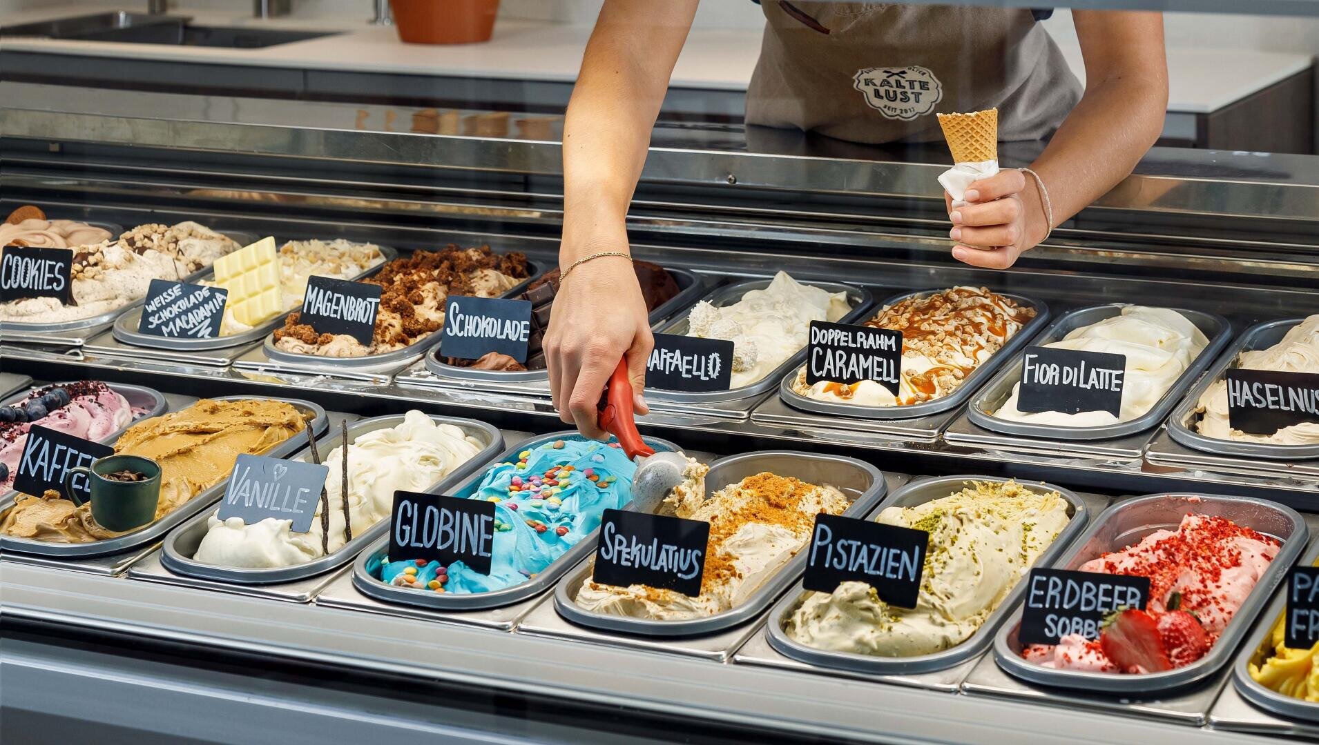 Eine Person schöpft blaues Eis aus einer Vitrine, die mit vielen bunten Gelato-Sorten gefüllt ist, die jeweils mit Schildern auf Deutsch beschriftet sind. Die Person hält eine Waffeltüte bereit, um das Eis zu servieren.