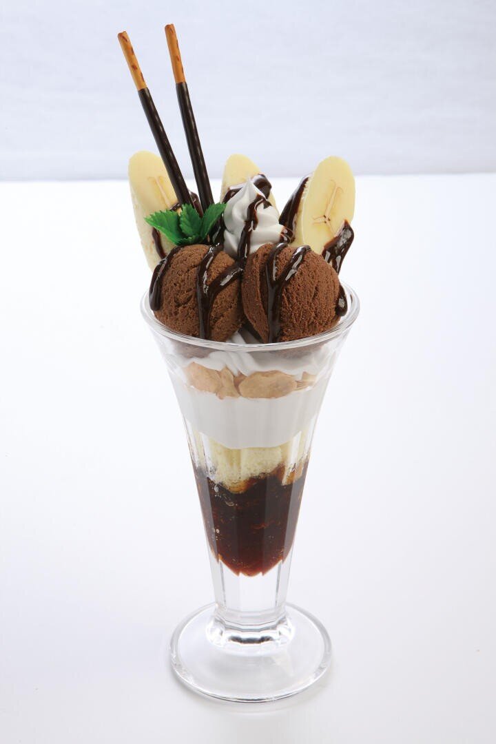 Ein Schokoladen-Bananen-Eisbecher in einem hohen Glas, garniert mit Kugeln Schokoladeneis, Bananenscheiben, Schlagsahne und mit Schokolade überzogenen Keksstangen.