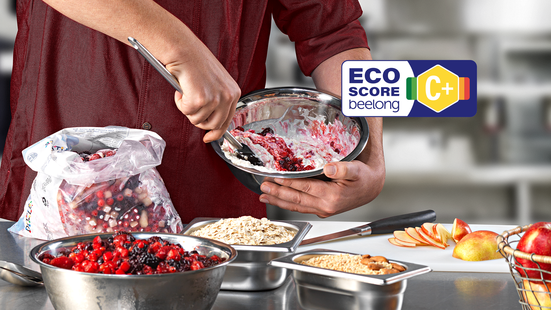 Eine Person mischt "Birchermüesli" Fruchtmischung und Joghurt in einer Schüssel. In der Nähe stehen Schüsseln mit Haferflocken, "Birchermüesli" Fruchtmischung und Apfelscheiben. Ein Eco-Score Label mit der Bewertung C+ zu sehen.
