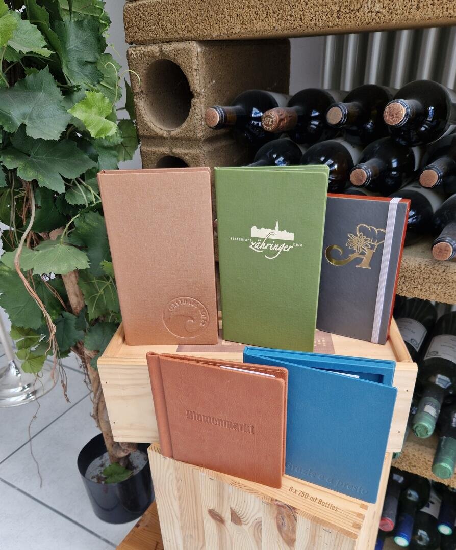 Eine Ausstellung von sechs farbenfrohen Weinkarten oder -tagebüchern, die aufrecht auf einer Holzkiste stehen, daneben Weinflaschen und grüne Weinblätter. Im Hintergrund sind ein Weinflaschenregal und eine Betonwand zu sehen.
