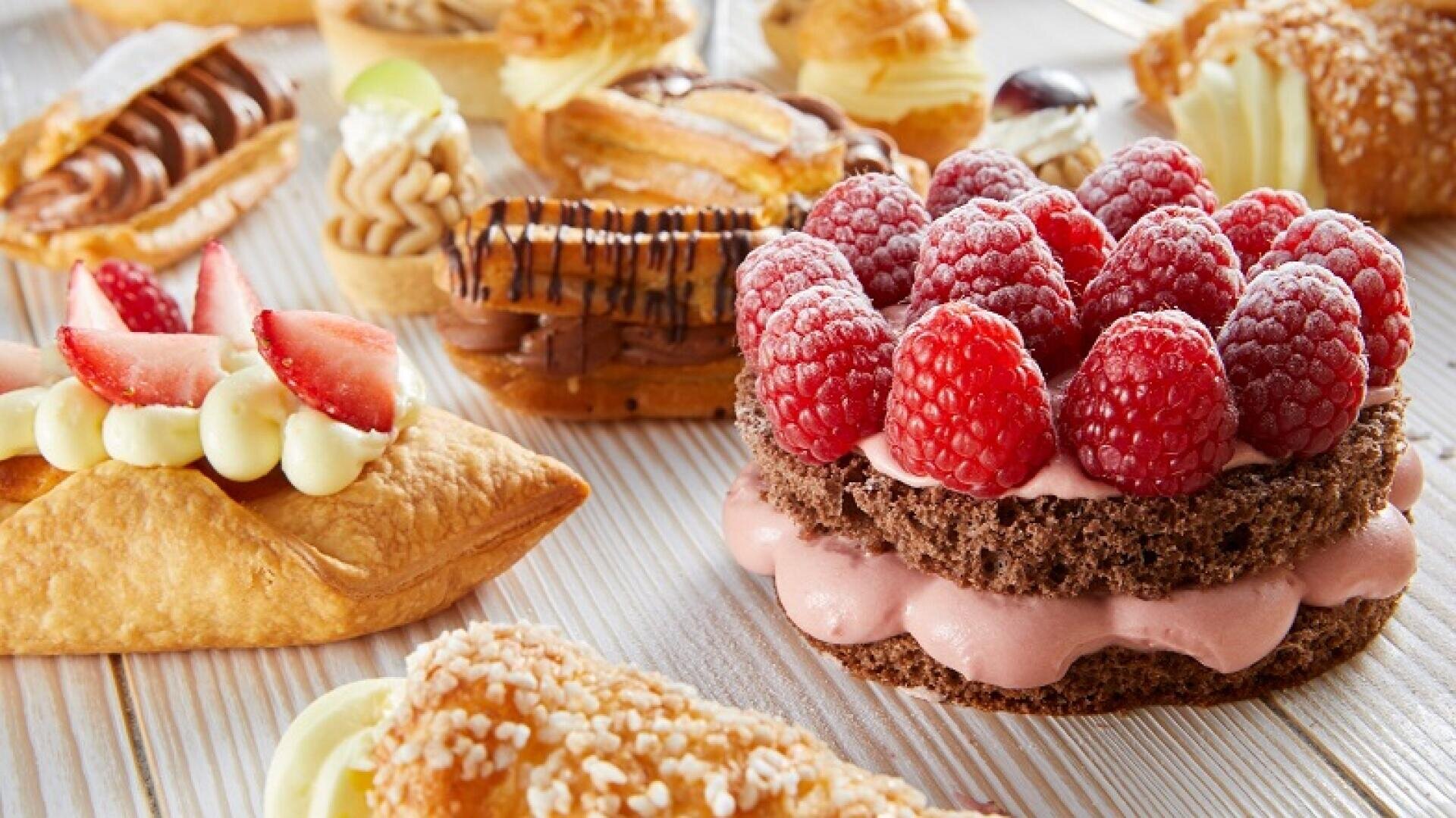 Eine Auswahl an Gebäck, darunter ein Schokoladen-Himbeer-Dessert, Obsttörtchen und mit Creme gefüllte Éclairs, präsentiert auf einer hellen Holzplatte.