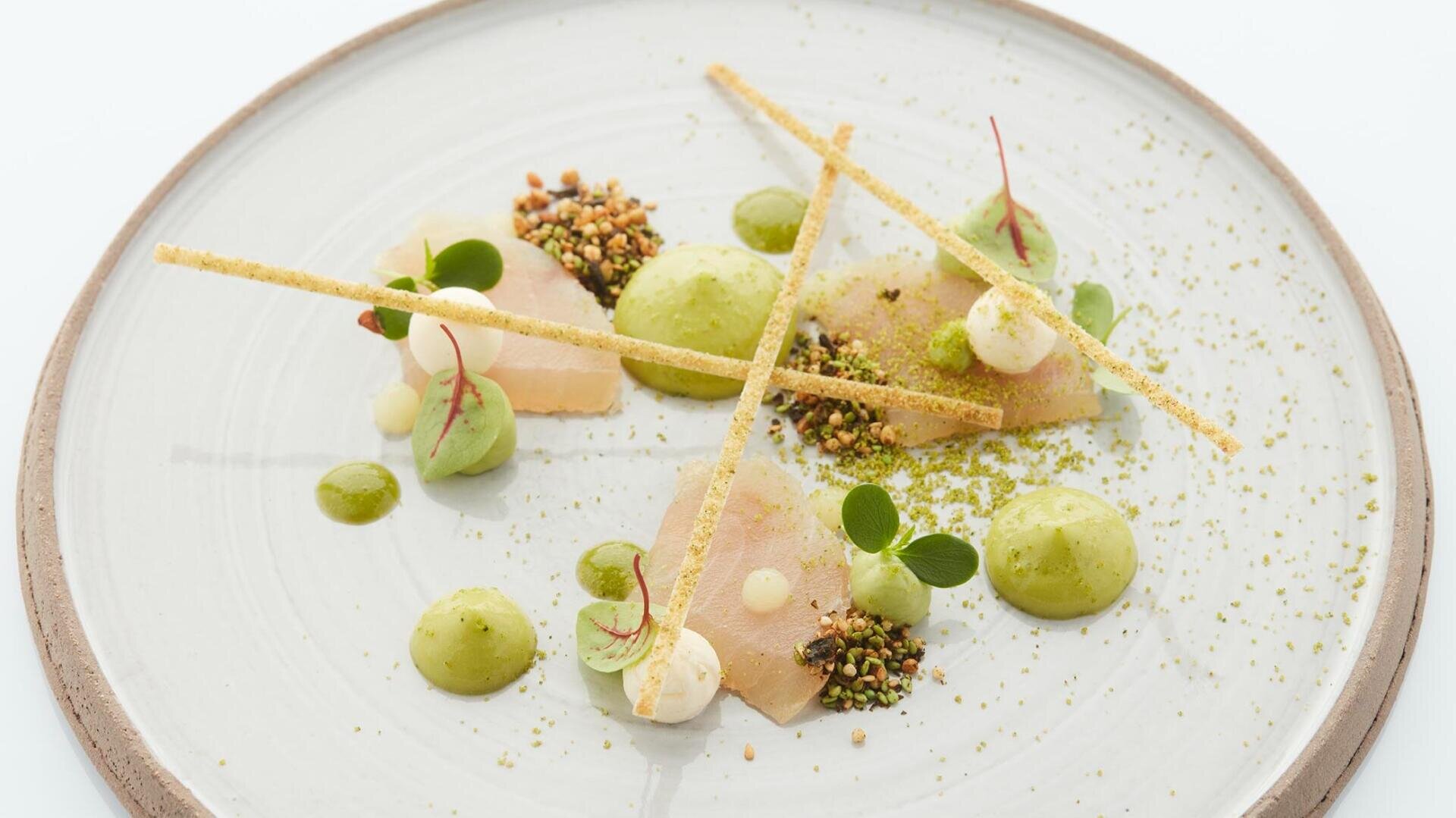 Kunstvoll angerichtetes Gourmetgericht mit dünnen Fischscheiben, grüner und weißer Soße, knusprigen Elementen und Microgreens, garniert mit dünnen, knusprigen Grissini auf einem weißen Teller mit einer Garnierung aus Pulver.