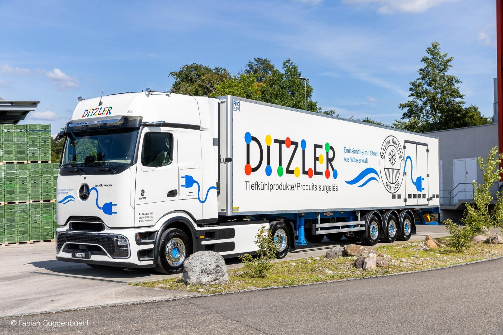 Ein weißer elektrischer Kühlwagen mit DITZLER-Schriftzug und blauen Steckern ist an einem sonnigen Tag vor einem Lagerhaus geparkt. In der Nähe sind grüne Kisten gestapelt, und im Hintergrund sind Bäume zu sehen.