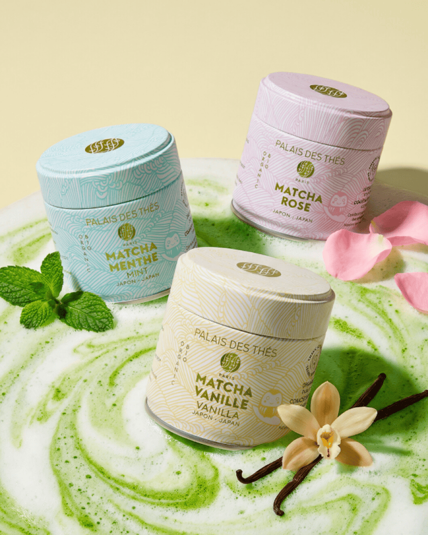 Drei pastellfarbene Teedosen mit den Aufschriften „Matcha Menthe“, „Matcha Rose“ und „Matcha Vanille“ stehen auf einer grün-weiß marmorierten Fläche, die mit Minzblättern, einer Vanilleblüte und einer rosa Blume verziert ist.