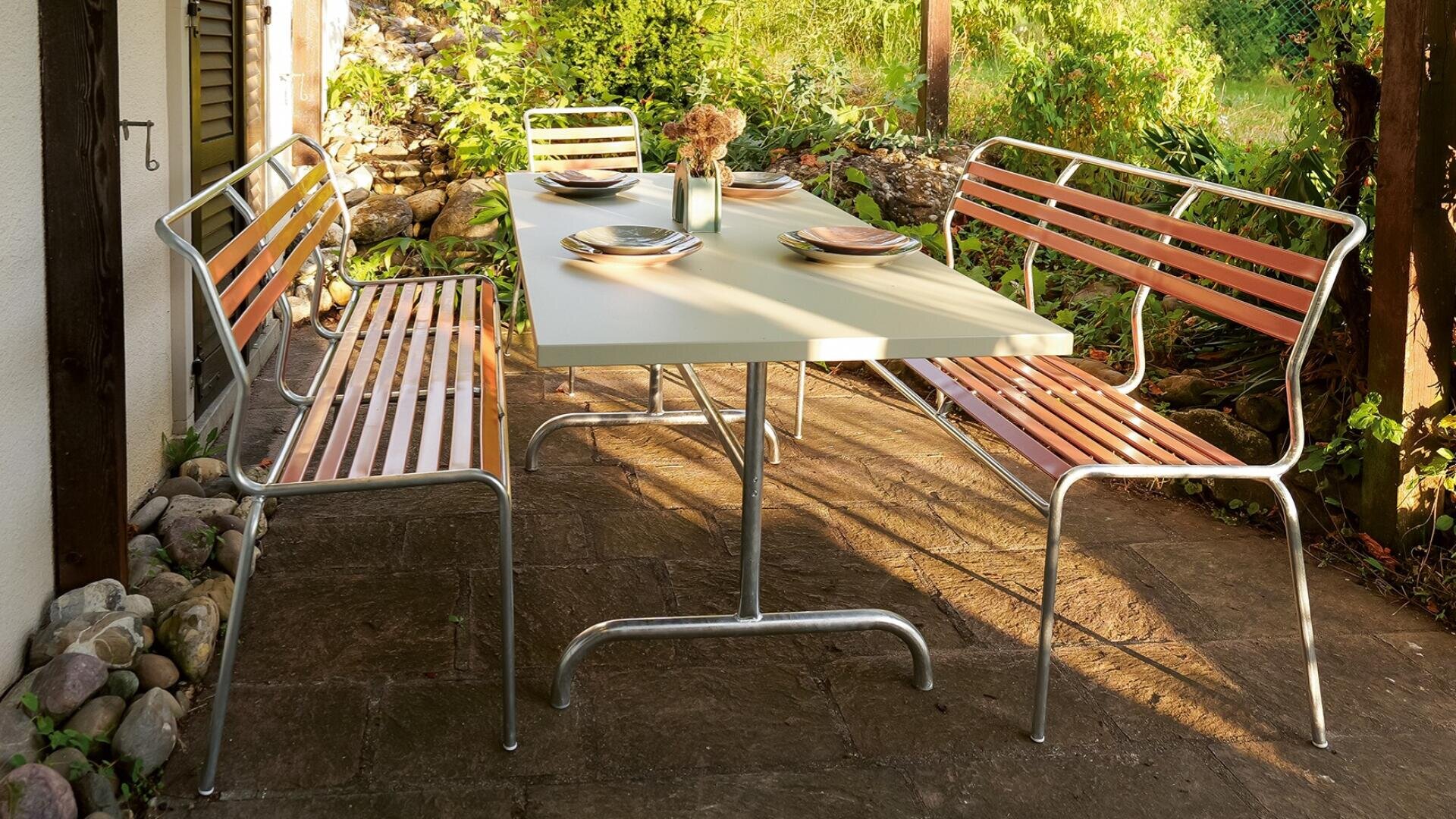 Eine Terrasse im Freien mit einem Metalltisch für vier Personen, flankiert von zwei Bänken aus Holzlatten. Umgeben von Grünpflanzen und Felsen wird die Szene vom Sonnenlicht erhellt, was eine gemütliche und einladende Atmosphäre schafft.