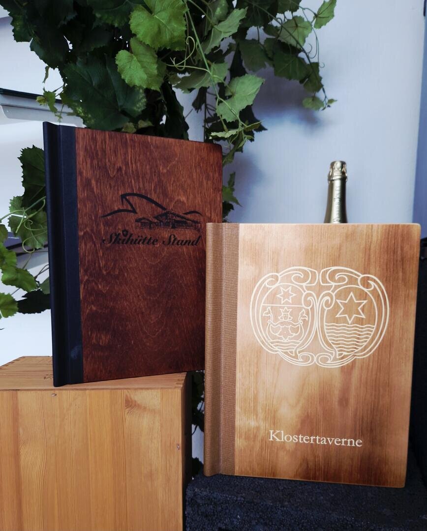 Neben einer Champagnerflasche und grünen Ranken stehen zwei Speisekartenhefte aus Holz: eines in dunkler Farbe mit der Aufschrift „Aphinitive Stand“ und einem Vogelmotiv, das andere in heller Farbe mit der Aufschrift „Klostertaverne“ und einem dekorativen Emblem.
