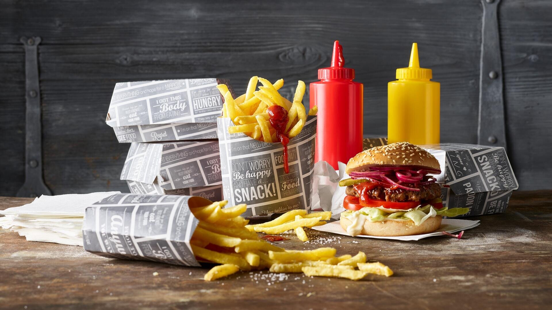 Auf einem rustikalen Holztisch liegt ein Burger mit Salat, Tomate, Zwiebel und Soße neben drei Portionen Pommes frites in Papierhüllen, im Hintergrund stehen Ketchup- und Senfflaschen.