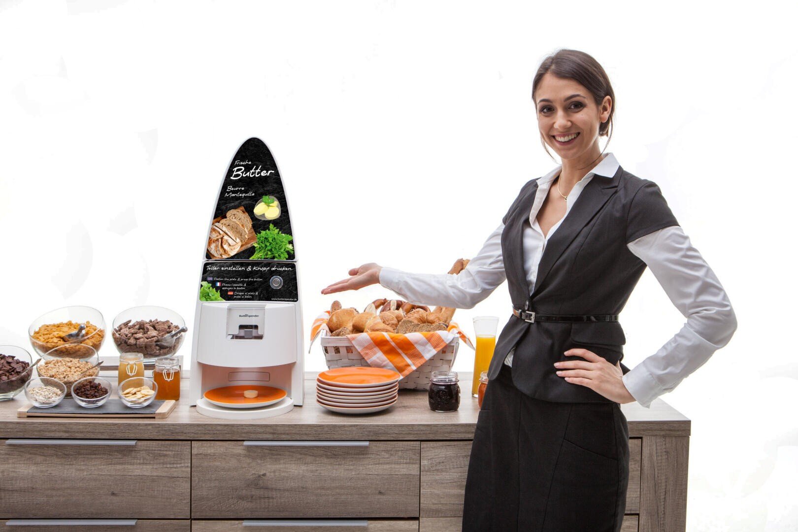 Eine lächelnde Frau in einer Weste deutet auf ein Frühstücksbuffet, auf dem ein automatischer Butterausgeber, Brot und Schalen mit Müsli stehen.