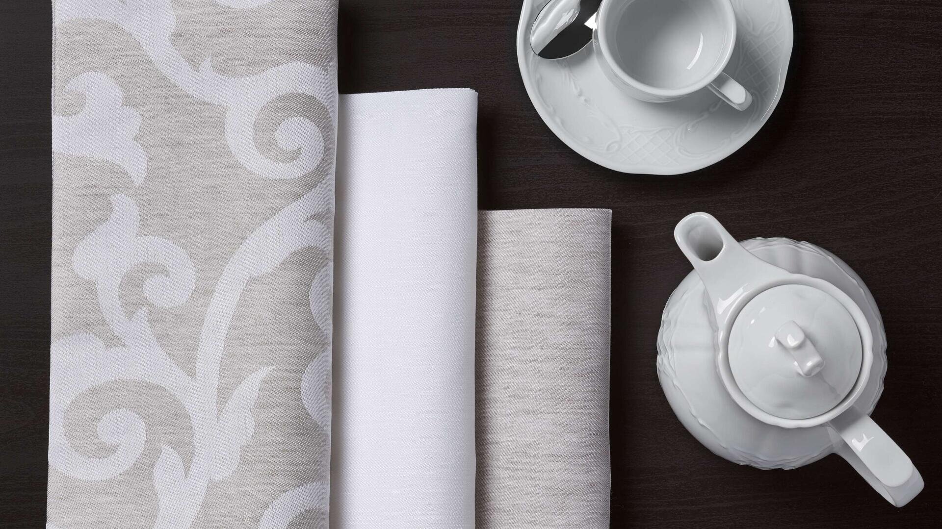Auf einer dunklen Unterlage liegen eine weiße Teekanne, eine Tasse mit Untertasse und Löffel sowie drei ordentlich gefaltete Tücher mit dezenten Mustern.