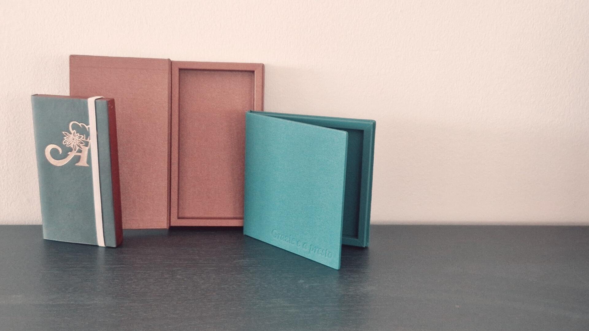 Drei Bücher stehen aufrecht auf einer dunklen Fläche vor einer hellen Wand: ein grünes Buch mit weißem Muster, ein rötliches, aufgeschlagenes Buch und ein aufgeschlagenes blaues Buch mit kaum lesbarem Text auf dem Einband.