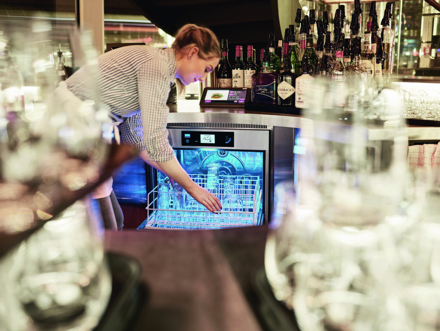 Eine Person stelht hinter einer Bar und nimmt saubere Gläser aus einer Spülmaschine der Marke MEIKO, Modell M-iClean U, im Vordergrund sind weitere saubere Gläser zu sehen.