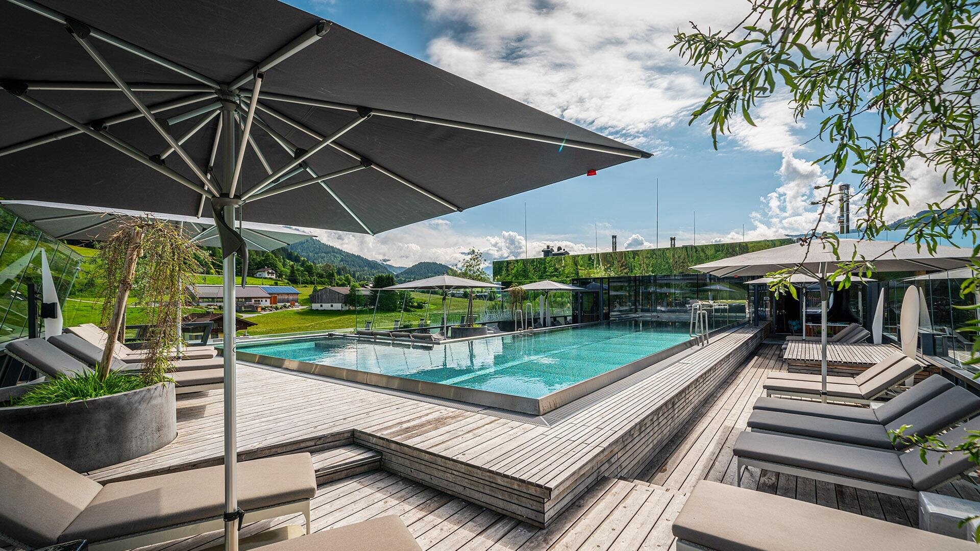 Moderner Außenpoolbereich mit Liegestühlen, großen Sonnenschirmen, Holzterrasse und malerischem Bergpanorama im Hintergrund unter einem teilweise bewölkten Himmel.