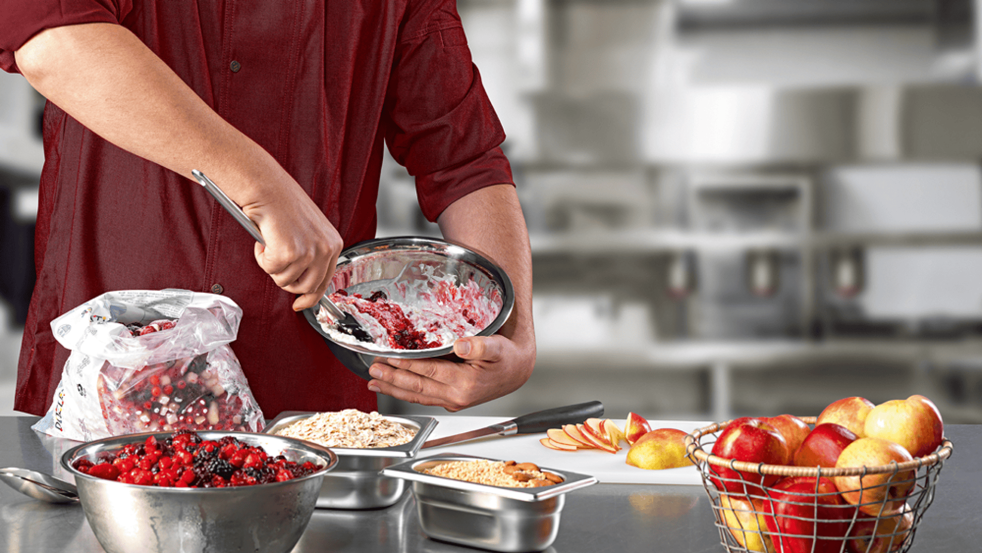 Eine Person in einem roten Hemd mischt Beeren und Haferflocken in einer Metallschüssel auf einem Küchentisch, in dessen Nähe Äpfel, Haferflocken, eine Beerenmischung und andere Zutaten angeordnet sind.