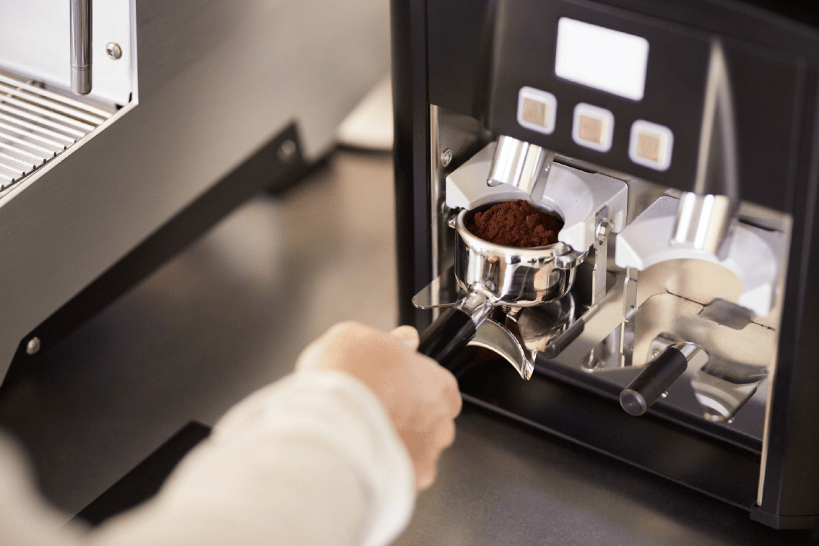 Eine Person hält einen mit gemahlenem Kaffee gefüllten Siebträger unter eine Maschine und bereitet vermutlich in einem Café oder in der Küche einen Espresso zu.