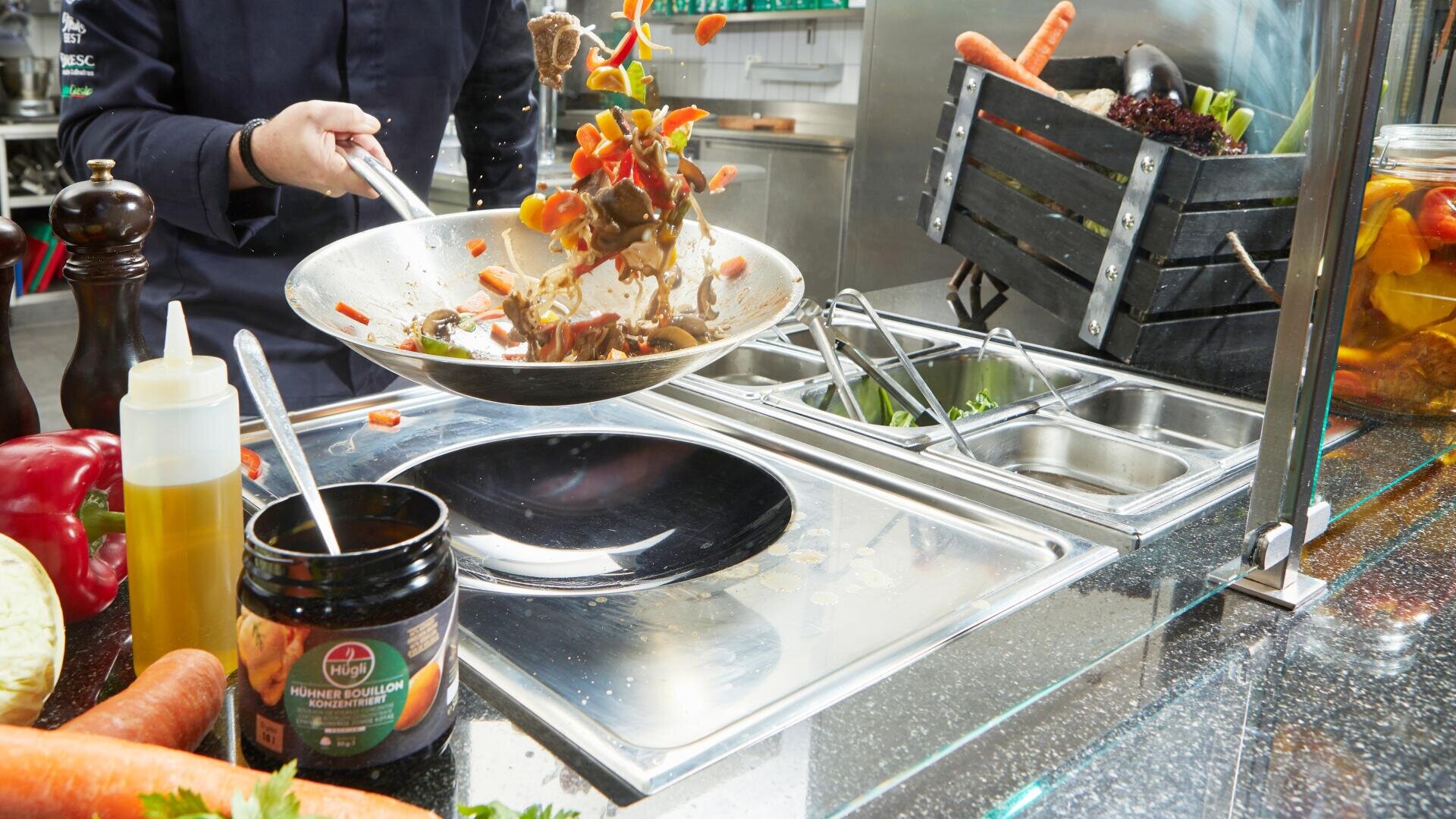 In einer hellen Küche schwingt ein Koch Gemüse in einer Pfanne über einer Arbeitsplatte, auf der Salatzutaten, Saucen und eine Holzkiste mit frischen Produkten stehen.