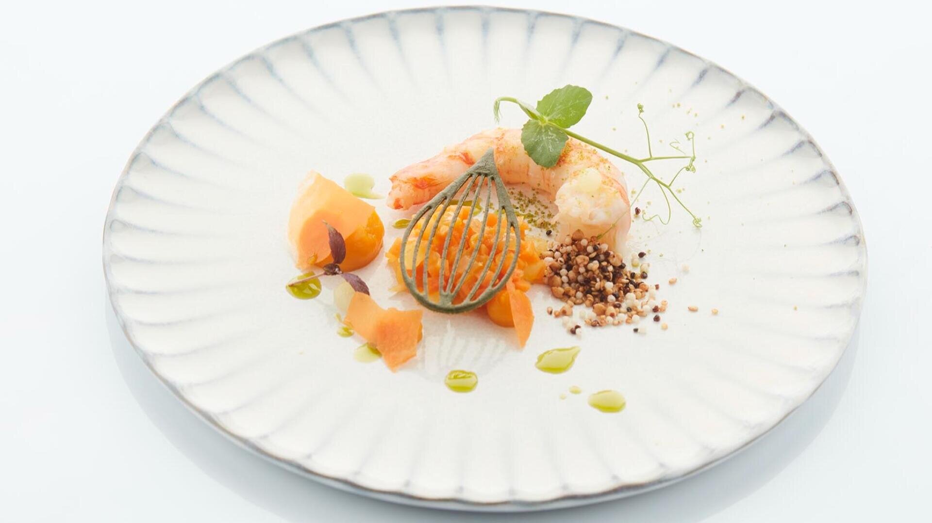 Ein Gourmetgericht mit Garnelen, orangefarbenen Gemüsescheiben, einer grünen Beilage, knusprigen Körnern und einem Spritzer grüner Soße, kunstvoll auf einem weißen, strukturierten Teller angerichtet.