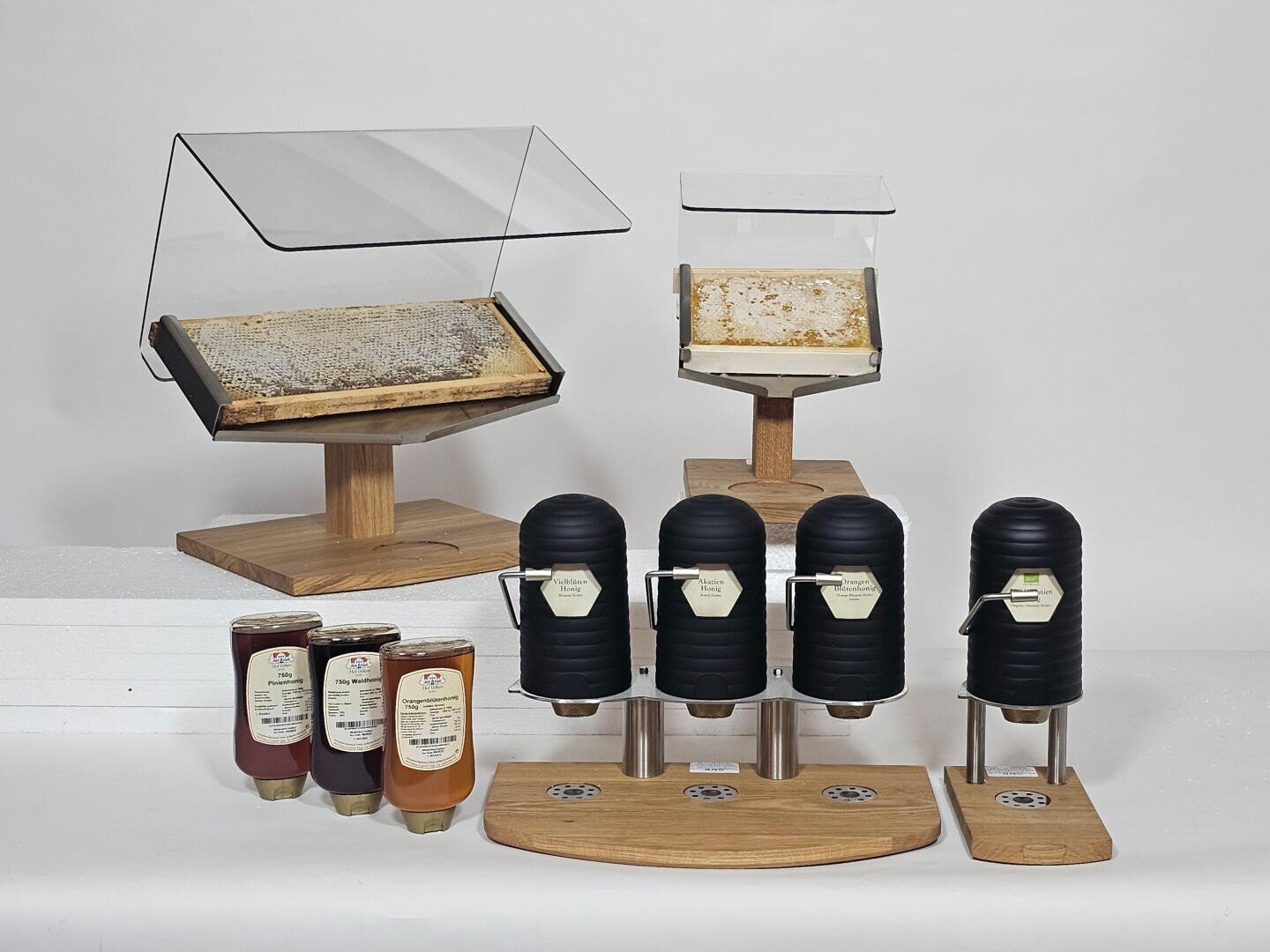 Zwei Waben auf Ausstellungsständern, drei Honigspender auf einem Holzsockel und drei Honigflaschen vor einem weißen Hintergrund, die zusammen eine Verkostungs- oder Servieranlage für Honig bilden.