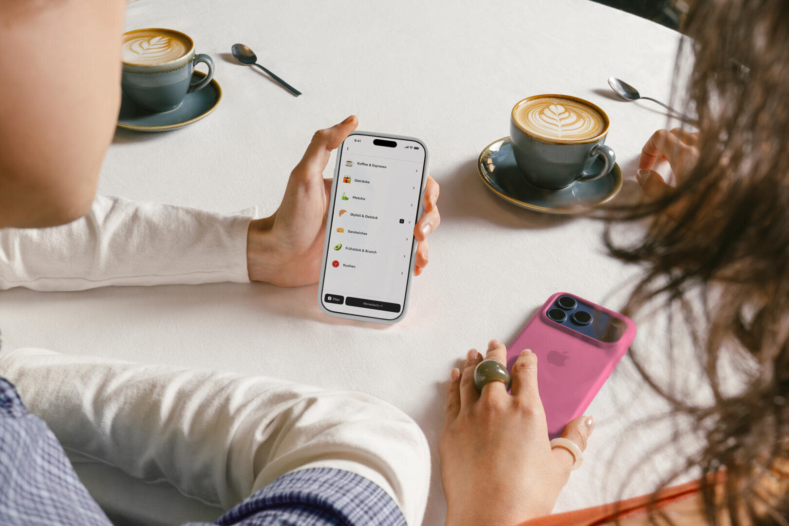 Zwei Personen sitzen an einem Cafétisch und schauen sich die Speisekarte auf einem Smartphone an; auf dem Tisch stehen zwei Tassen Latte und ein weiteres Smartphone in einer rosa Hülle.